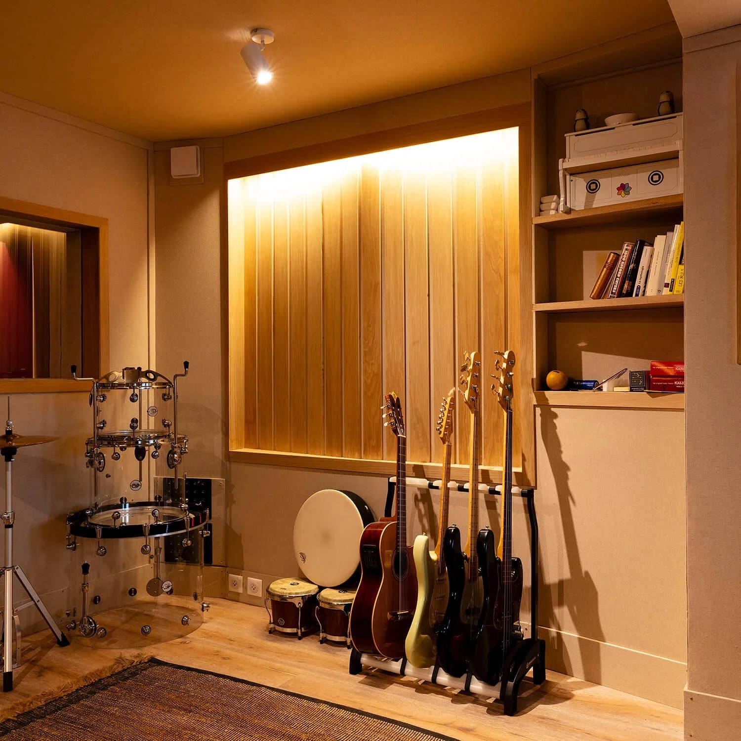 Une pièce de musique avec des guitares, une percussion, une étagère avec des livres, des boîtes et des objets. Mur en bois et sol en parquet.