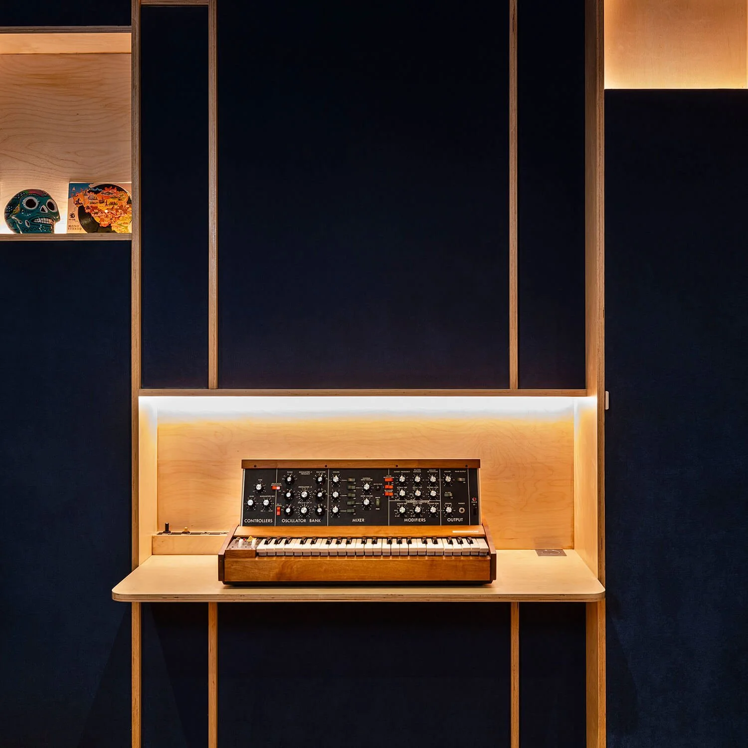 Clavier synthétiseur vintage en bois dans un meuble en bois clair, avec éclairage intégré, contre un mur noir.