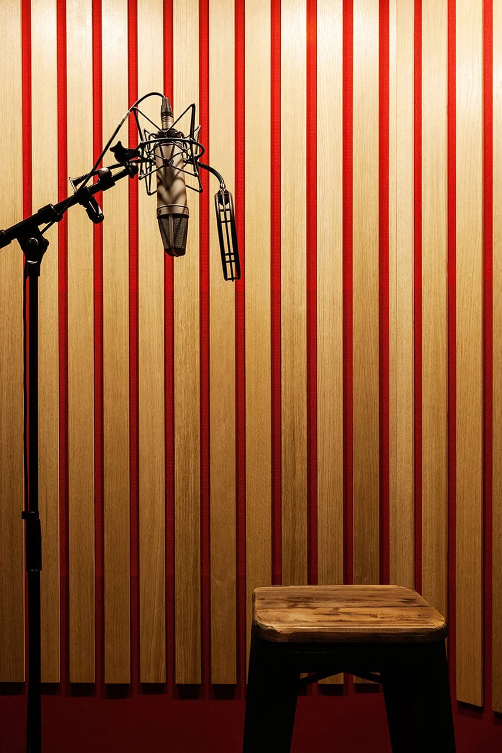 Une cabine d'enregistrement avec un microphone suspendu et un tabouret en bois, murs en bois clair avec rayures rouges.