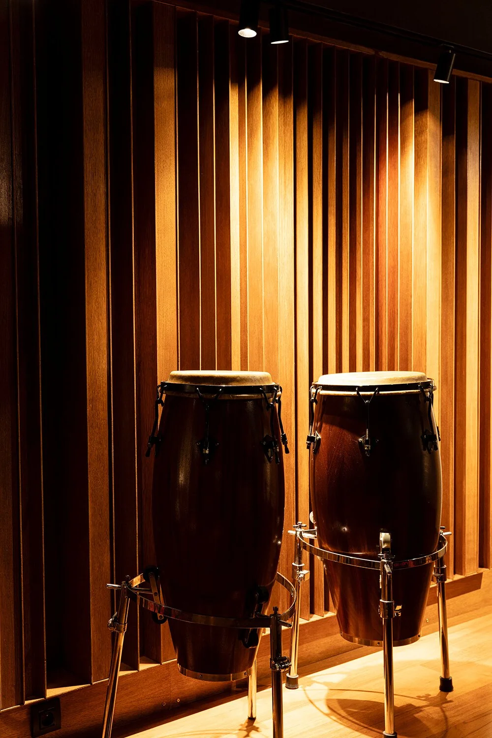 Deux congas en bois coloré, placées contre un mur en bois avec des rayons obliques, éclairées par un projecteur au plafond.