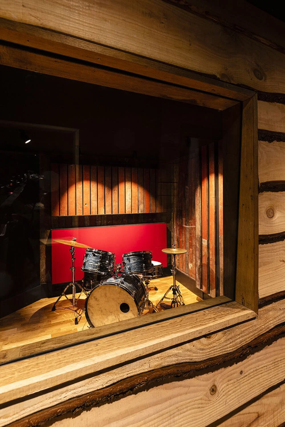 Une batterie de musique vue à travers une fenêtre dans un cadre en bois dans une salle de studio ou de répétition, avec un mur en bois derrière.