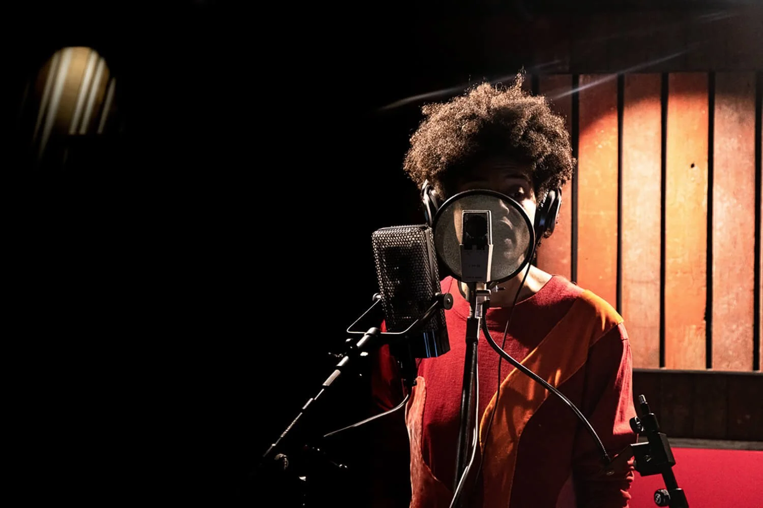 Une personne dans un studio d'enregistrement, portant un casque audio, chantant devant un microphone dans une pièce sombre avec un éclairage chaleureux.