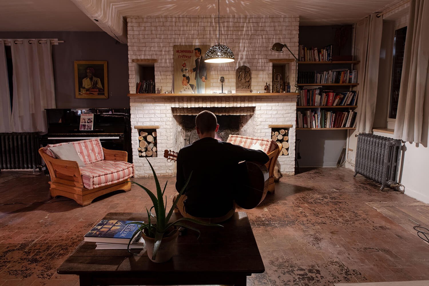 Une personne jouant de la guitare dans un salon avec cheminée en briques blanches, livres sur des étagères, un piano noir, une plante en pot sur une table et une lampe suspendue.