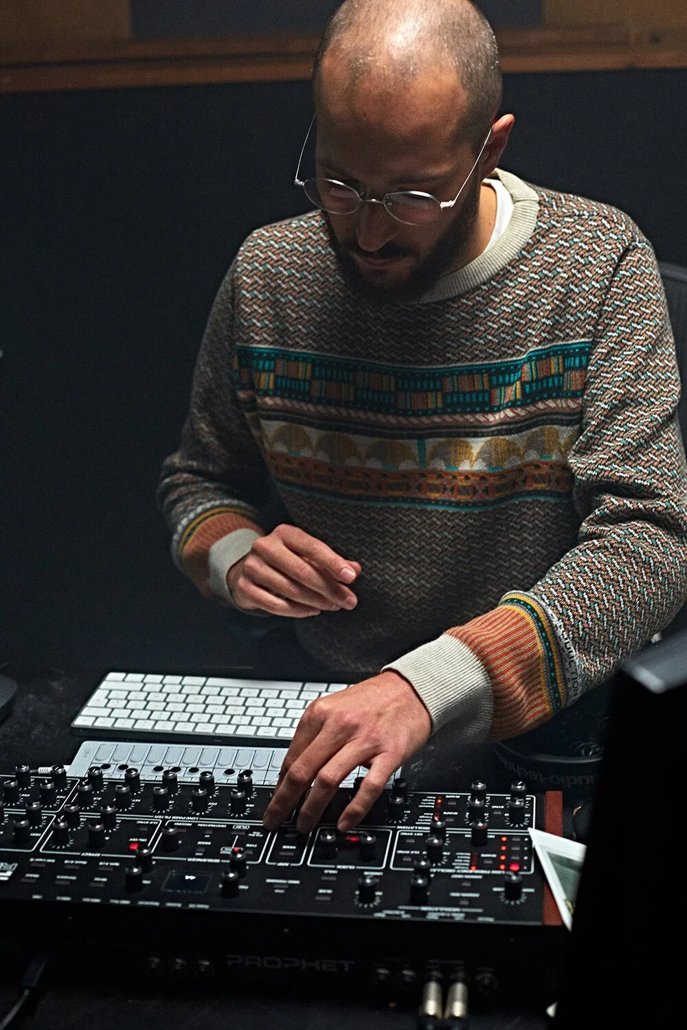Un homme avec des lunettes, portant un pull à motifs, manipulant un équipement de musique dans une ambiance sombre.
