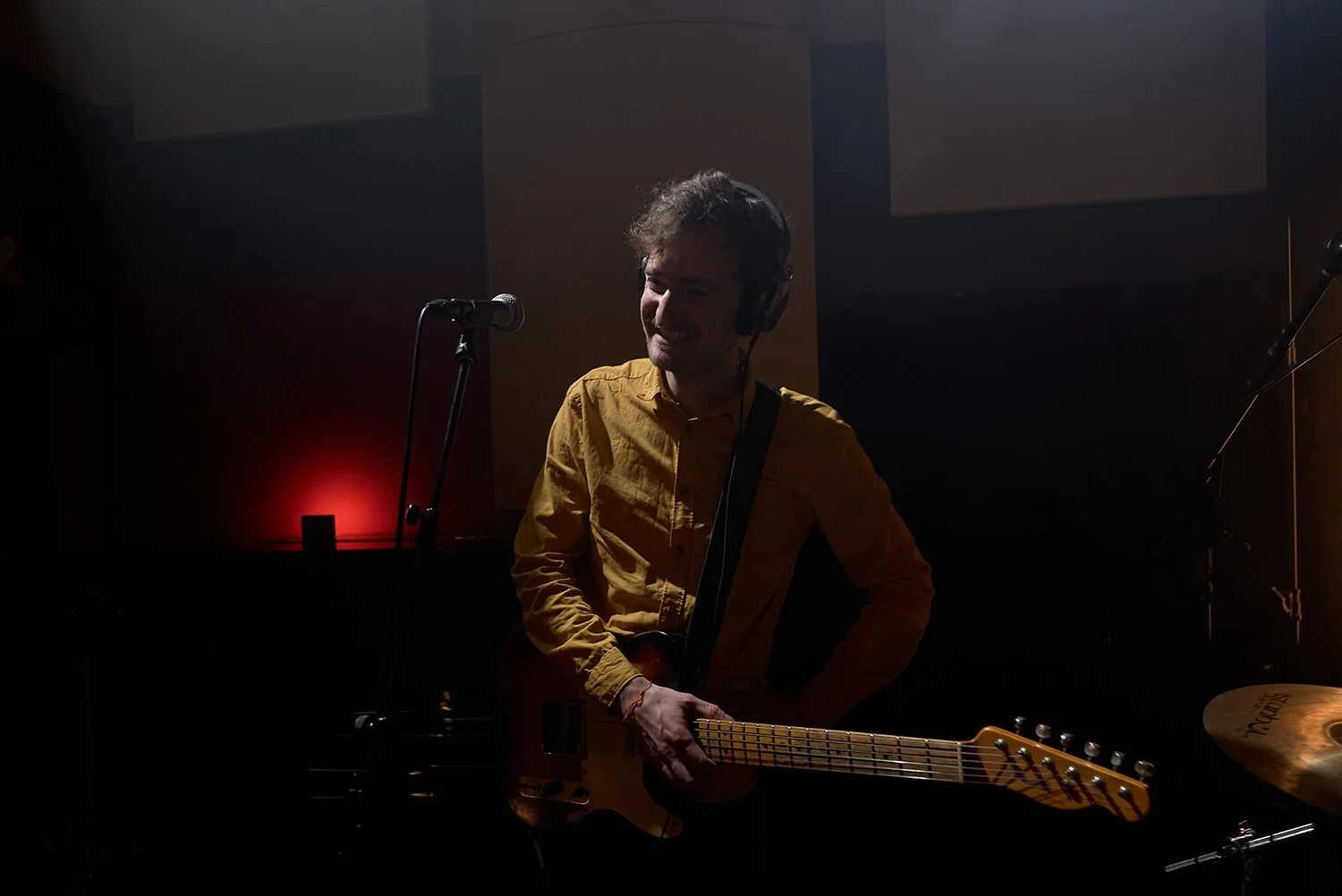 Un musicien souriant portant une chemise jaune, tenant une guitare électrique, dans une pièce sombre avec un micro et un kit de batterie visible.