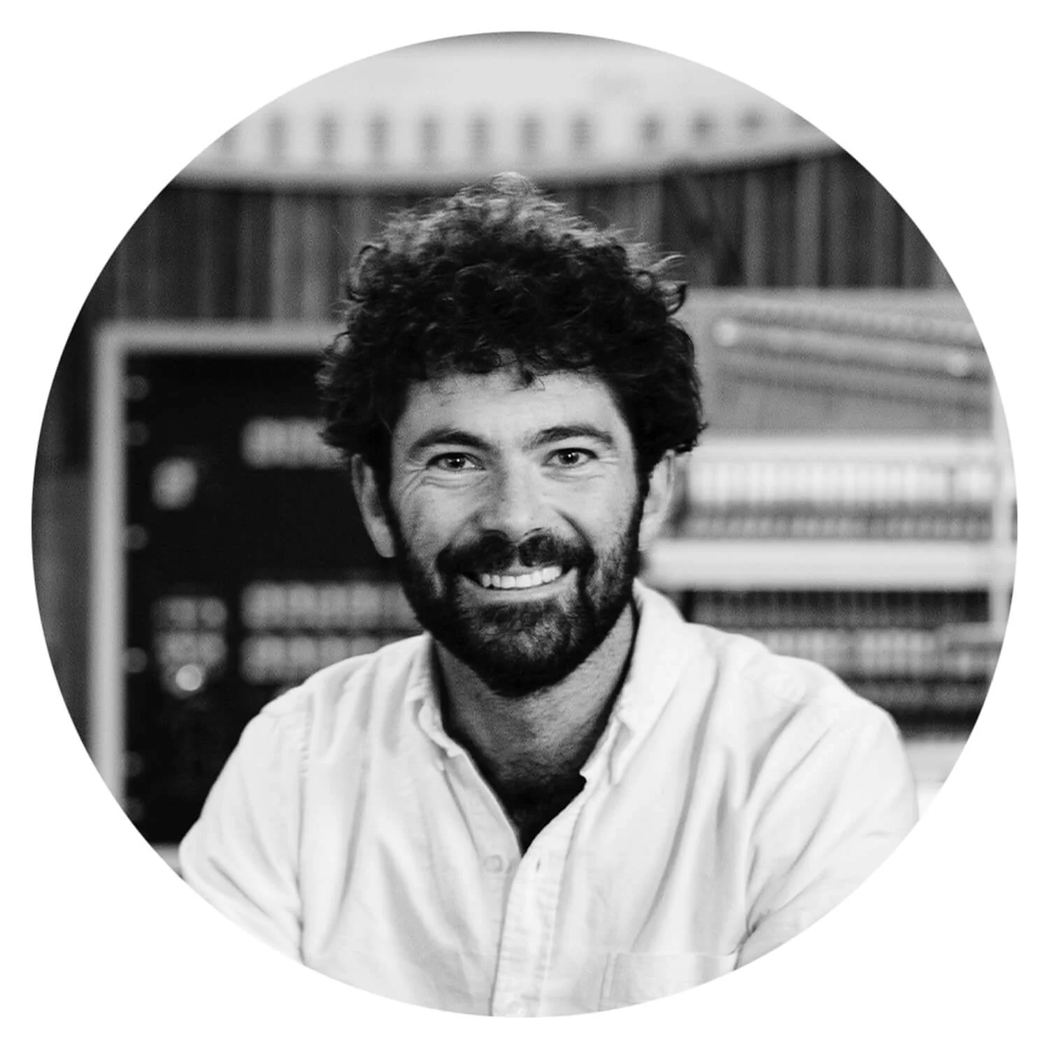 Homme souriant avec barbe et cheveux bouclés, portant une chemise blanche, en bibliothèque ou bureau avec des étagères remplies de livres en arrière-plan.