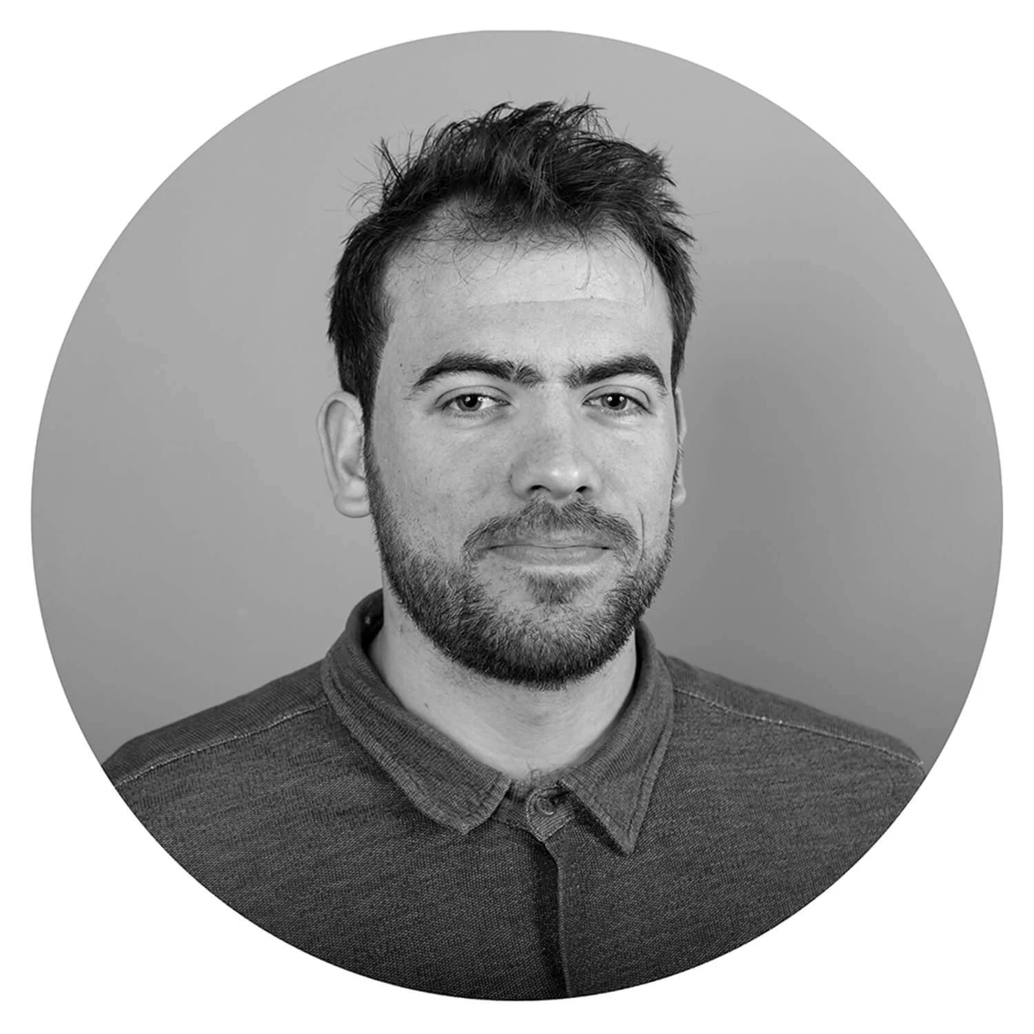 Portrait en noir et blanc d'un homme avec une barbe, cheveux foncés et veston à col. Fond gris clair.