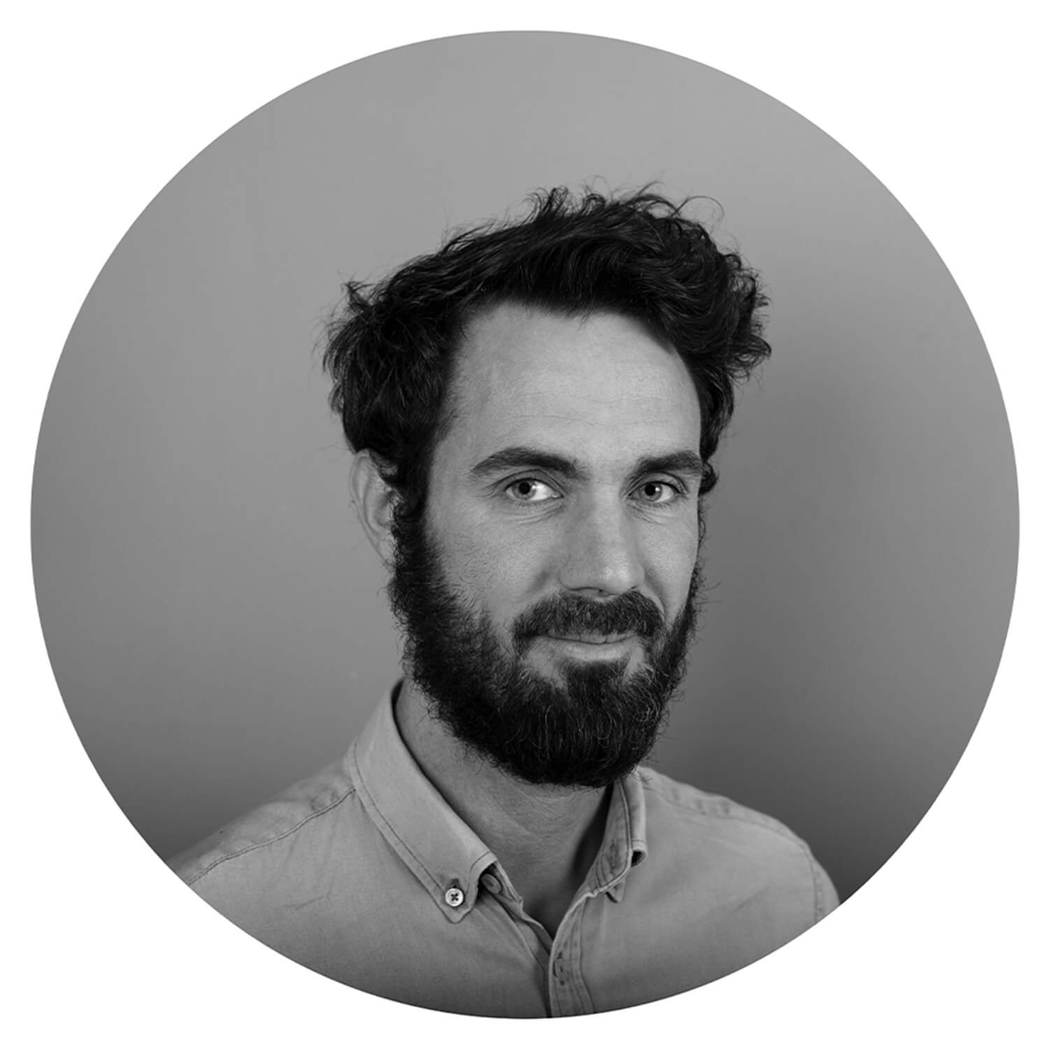 Photo en noir et blanc d'un homme avec une barbe et des cheveux bouclés, portant une chemise, regardant vers l'objectif, fond uni gris