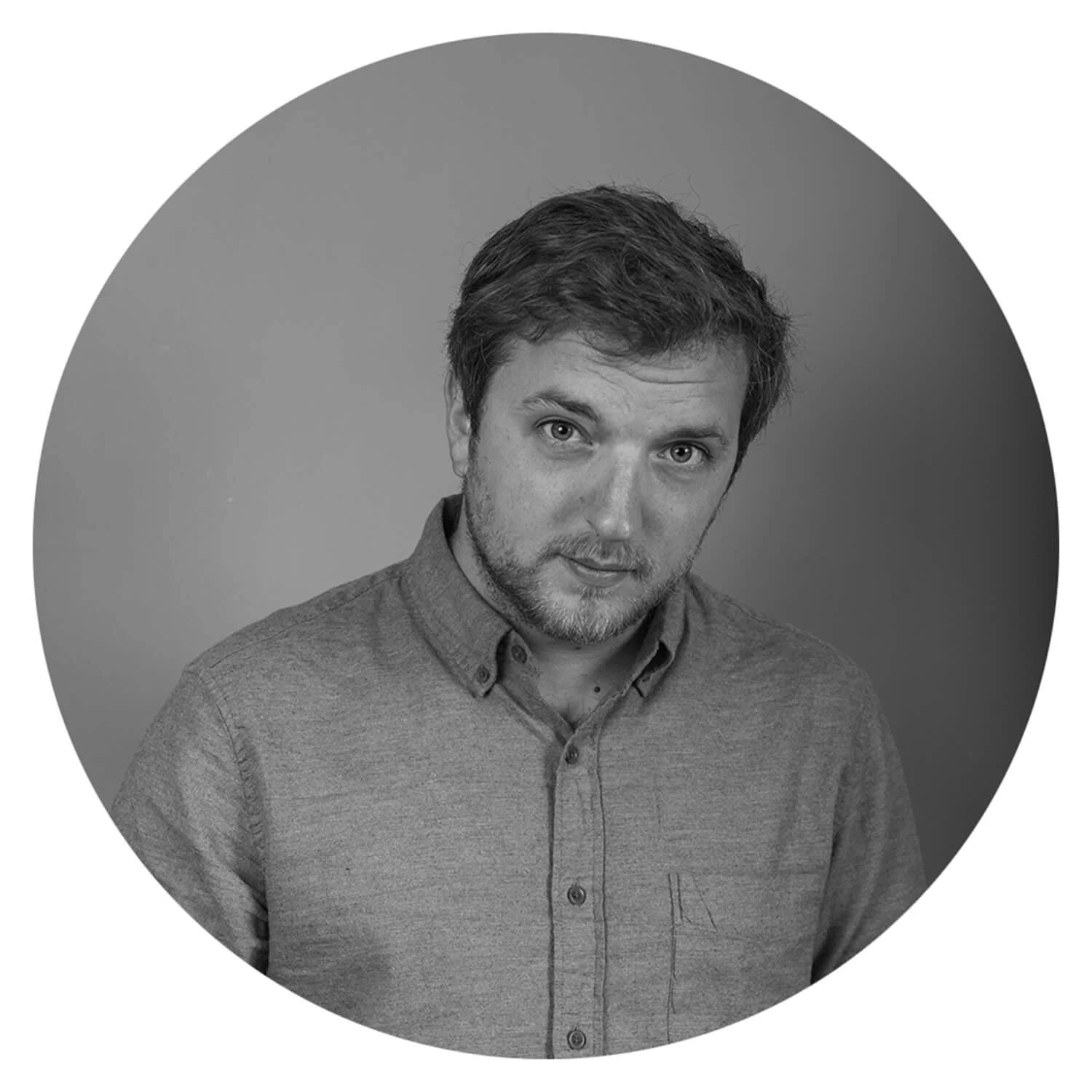 Photo en noir et blanc d'un homme avec des cheveux courts, une barbe légère, portant une chemise à boutons, regardant l'objectif dans un fond gris clair.