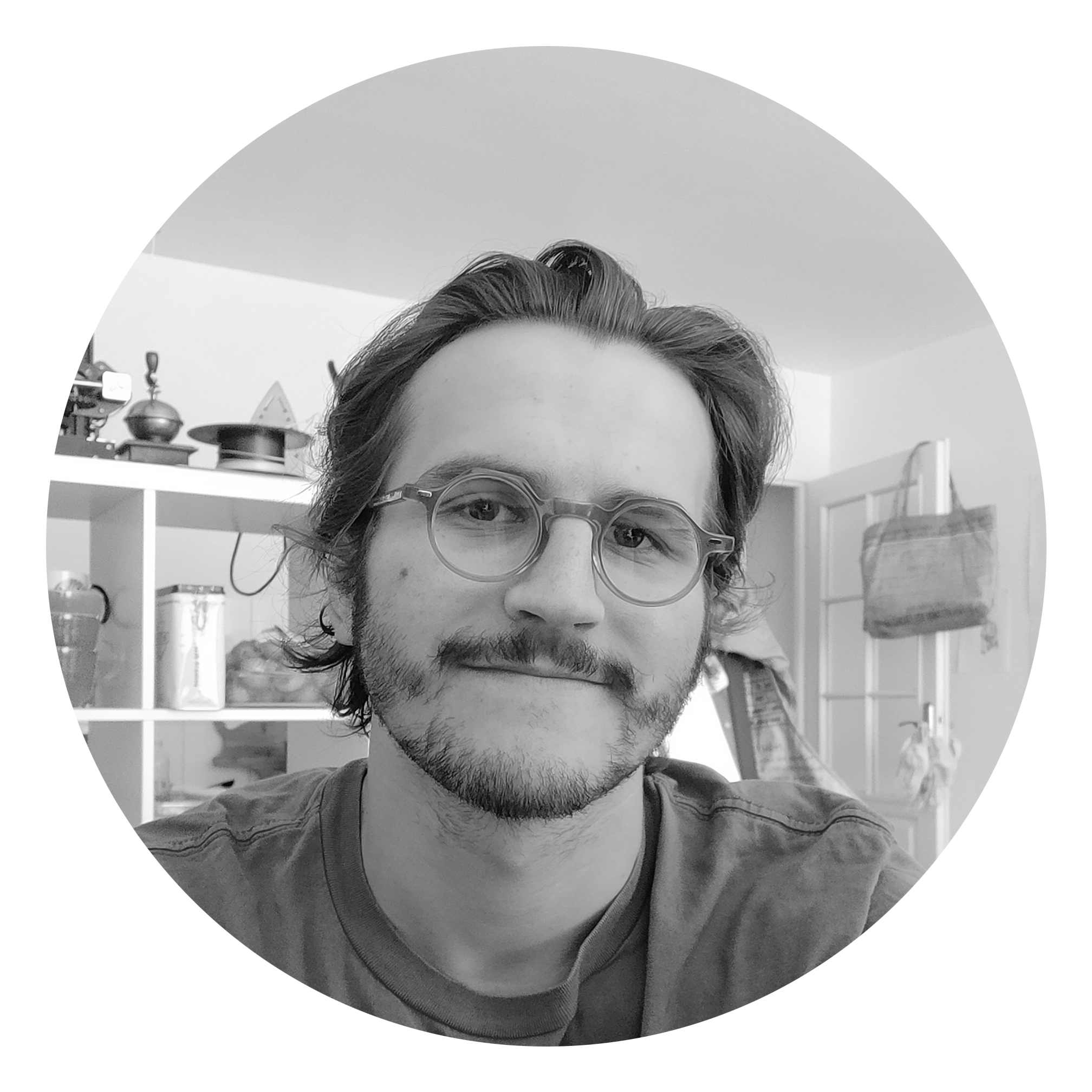 Un homme avec des lunettes, barbe et cheveux longs, regarde la caméra, dans une pièce avec étagères derrière lui contenant des objets divers.