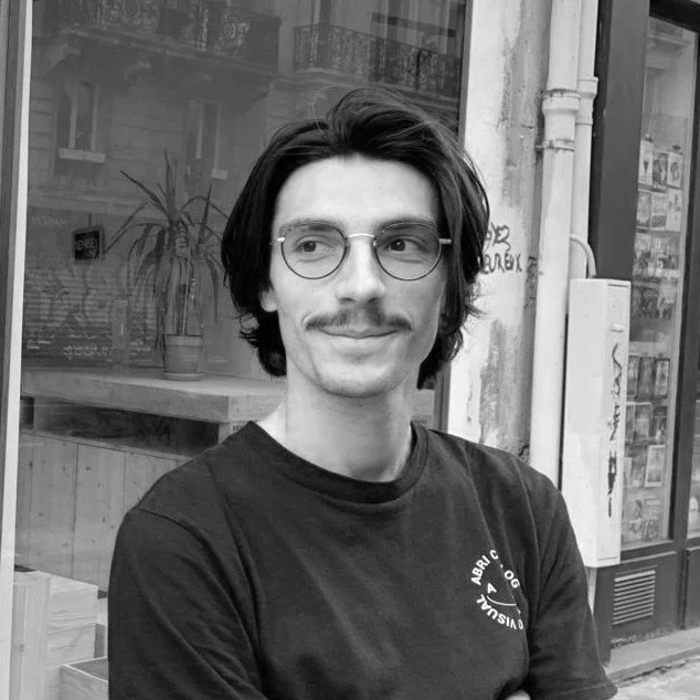 Jeune homme avec des lunettes, moustache, souriant, portant un t-shirt noir, assis en extérieur, arrière-plan avec des plantes et une fenêtre.