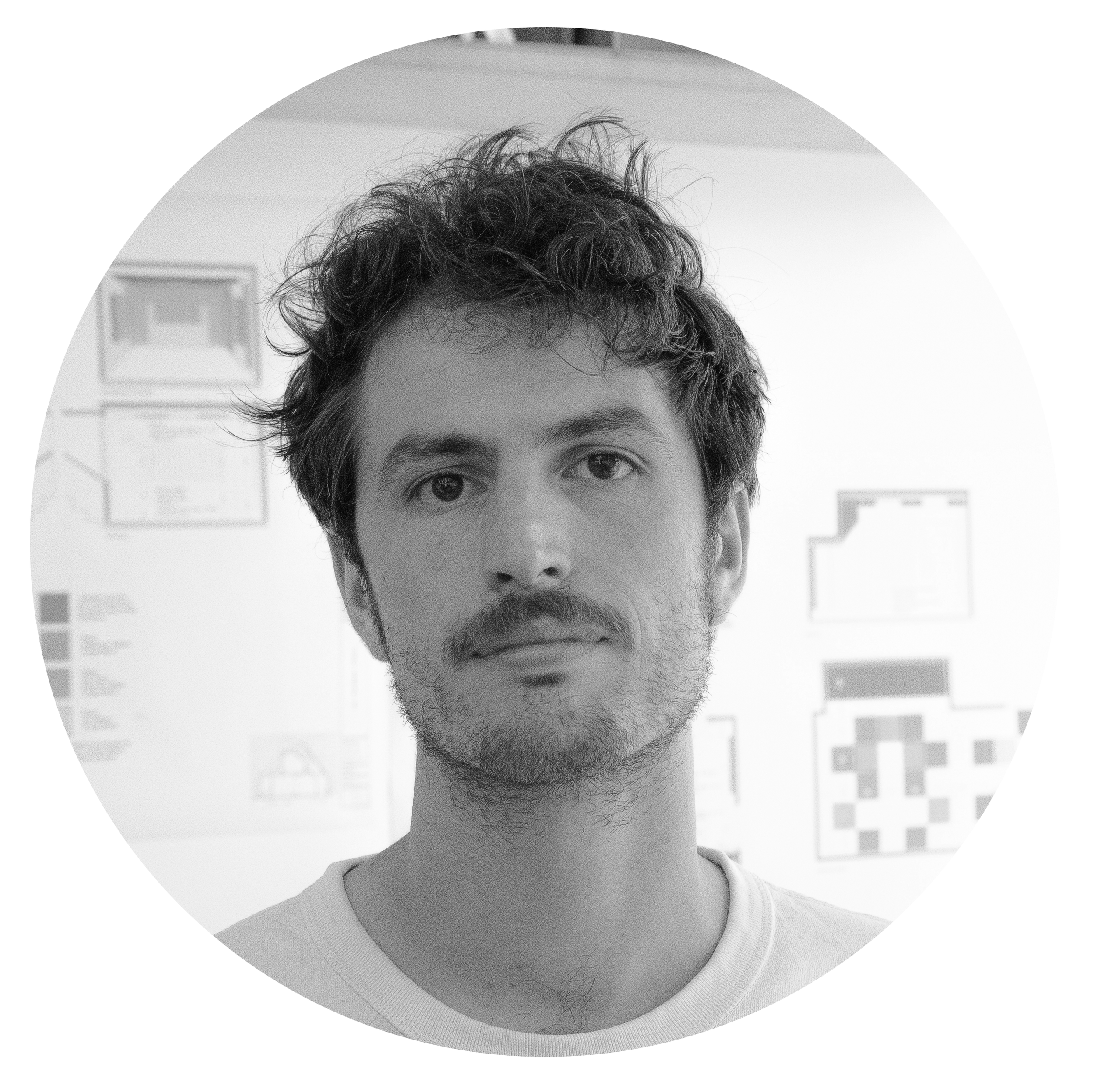 Un jeune homme avec des cheveux bouclés et une barbe, regardant l'objectif, en arrière-plan des plans et des schémas affichés sur un mur.