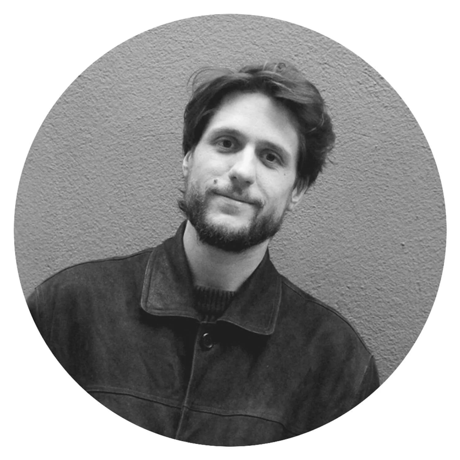 Un homme avec une barbe, portant une veste en jean noire, sourit légèrement, posé devant un mur texturé.