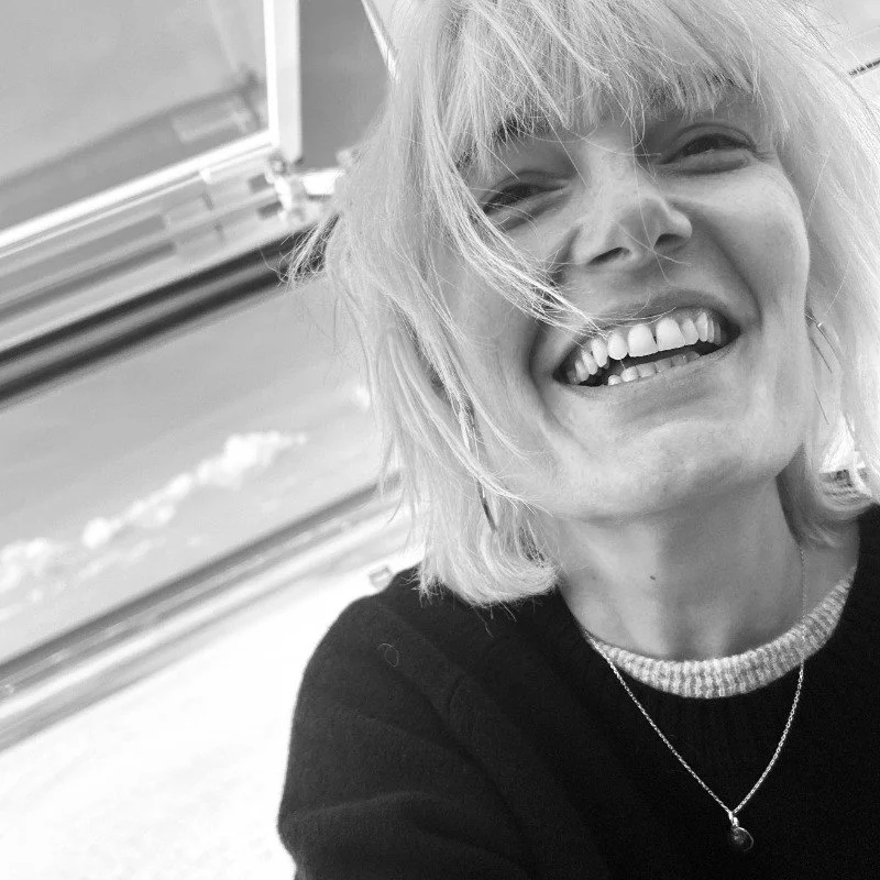 Une femme souriante avec des cheveux blonds, portant un pull noir et un collier, photo en noir et blanc.