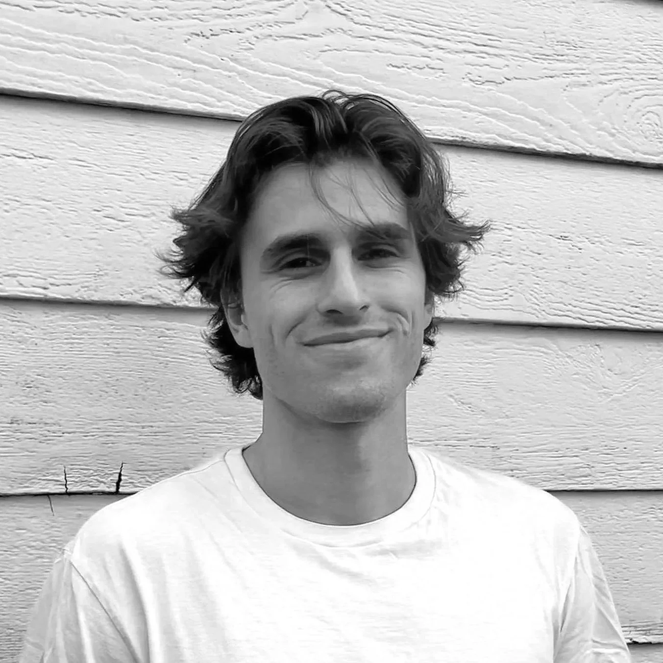 Un jeune homme souriant avec des cheveux bouclés, portant un t-shirt blanc, se tient devant un mur en bois.