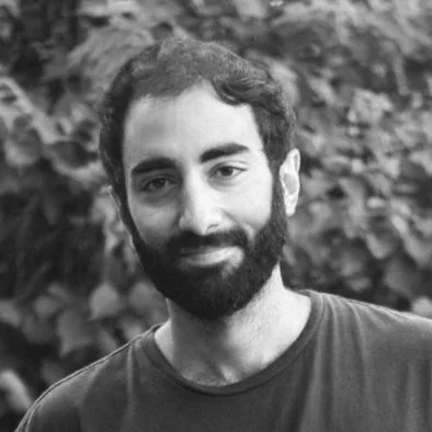 Un homme avec une barbe, souriant, en face d'un fond végétal.