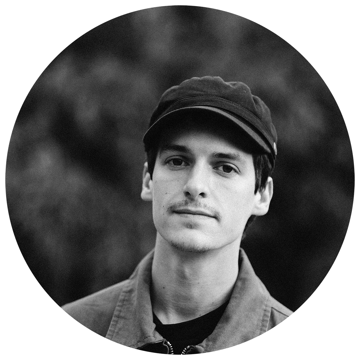 Un jeune homme portant une casquette et une veste en jean, regardant vers l'objectif, en noir et blanc.