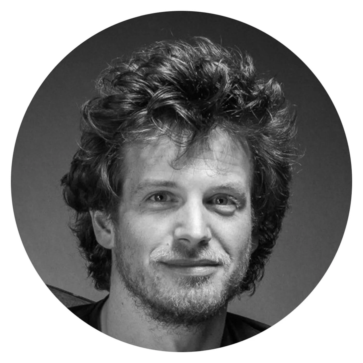 Portrait noir et blanc d'un homme avec des cheveux bouclés et une barbe légère, souriant légèrement.