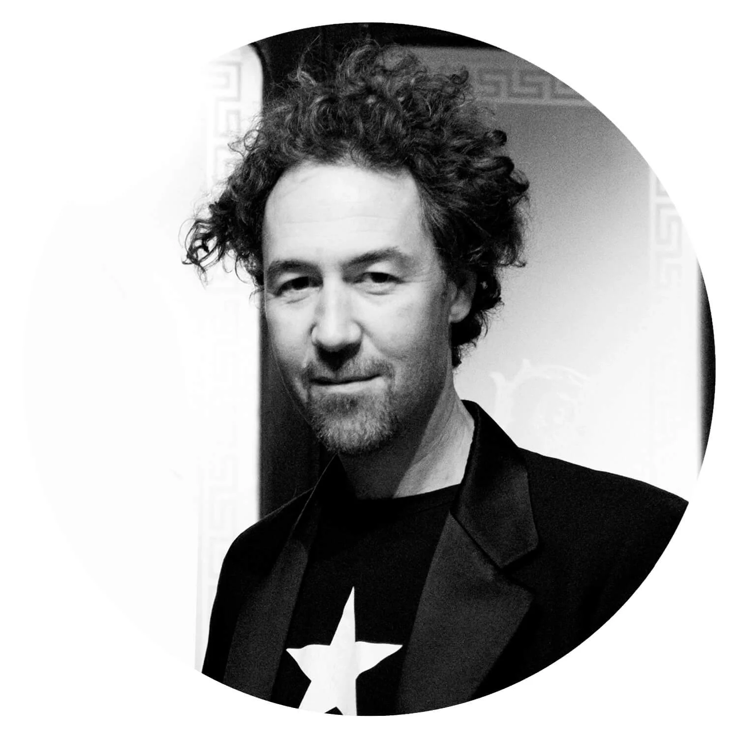 Portrait d'un homme avec des cheveux bouclés, portant un blazer noir et un t-shirt avec une étoile blanche, en noir et blanc.