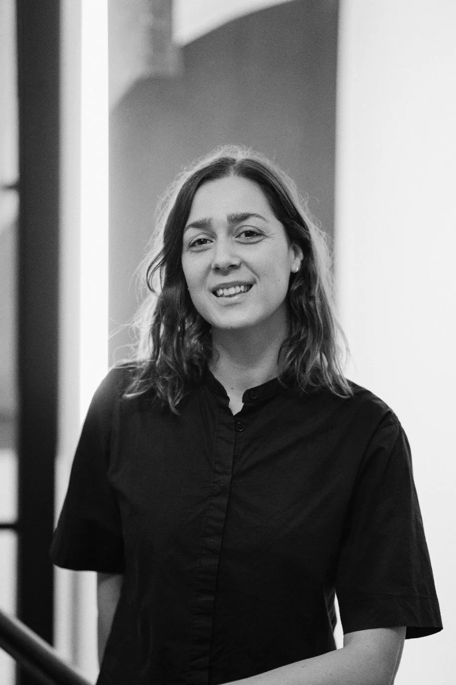 Portrait noir et blanc d'une femme souriante avec des cheveux ondulés, portant une chemise sombre, dans un intérieur moderne.