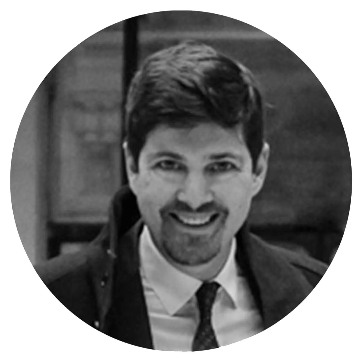 Un homme souriant, portant une veste sombre, une chemise blanche et une cravate, dans un style noir et blanc.