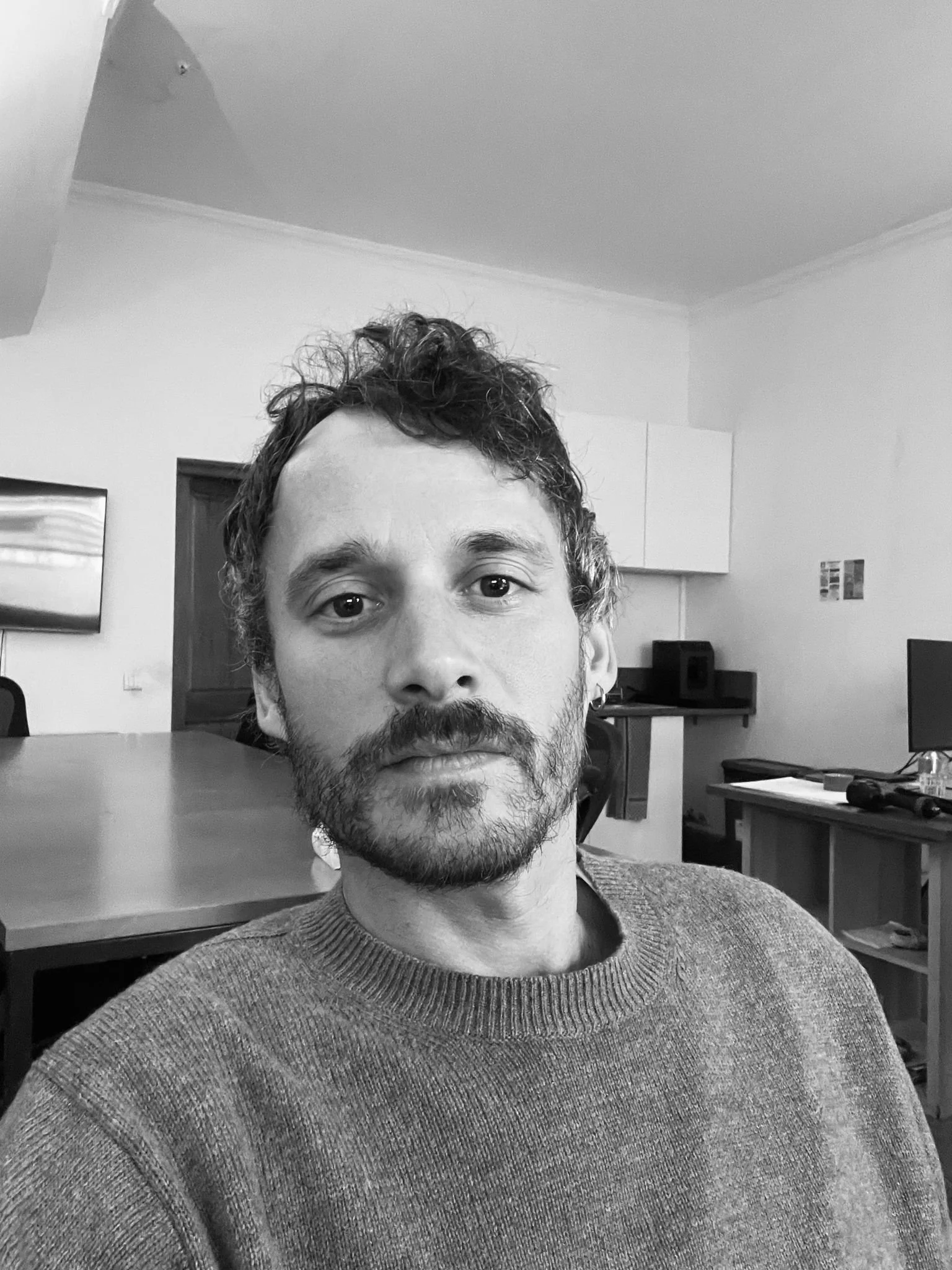 Portrait d'un homme avec barbe et cheveux bouclés dans une pièce avec meubles et écrans.