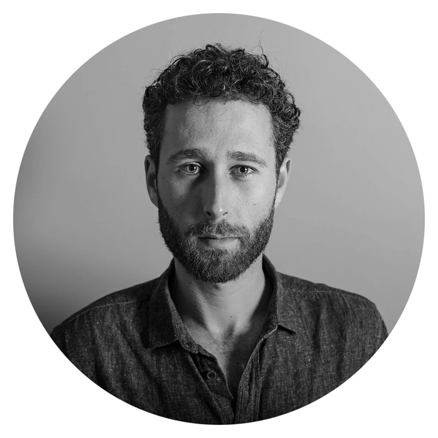 Photo en noir et blanc d'un homme avec des cheveux bouclés et une barbe, portant une chemise en denim, regardant directement l'objectif.