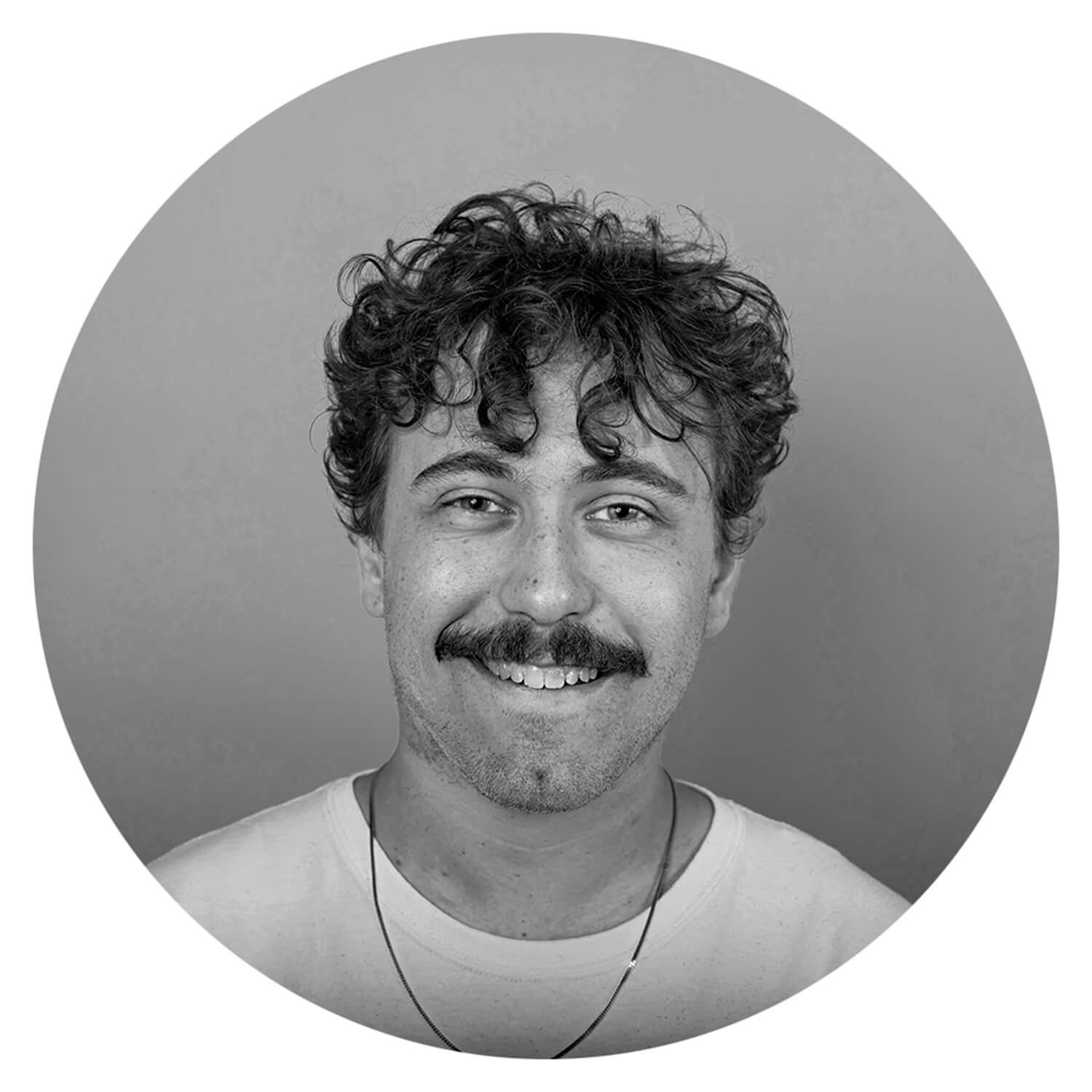 Portrait en noir et blanc d'un homme souriant avec des cheveux bouclés et une moustache, portant un T-shirt clair et une chaîne.