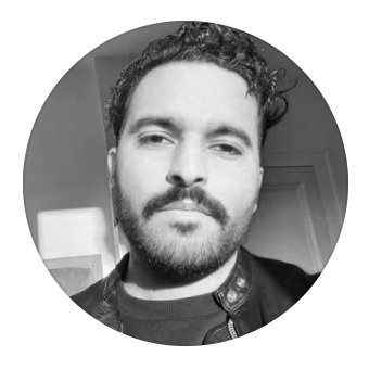 Un homme avec une barbe et des cheveux courts, regardant vers l'objectif, en noir et blanc.