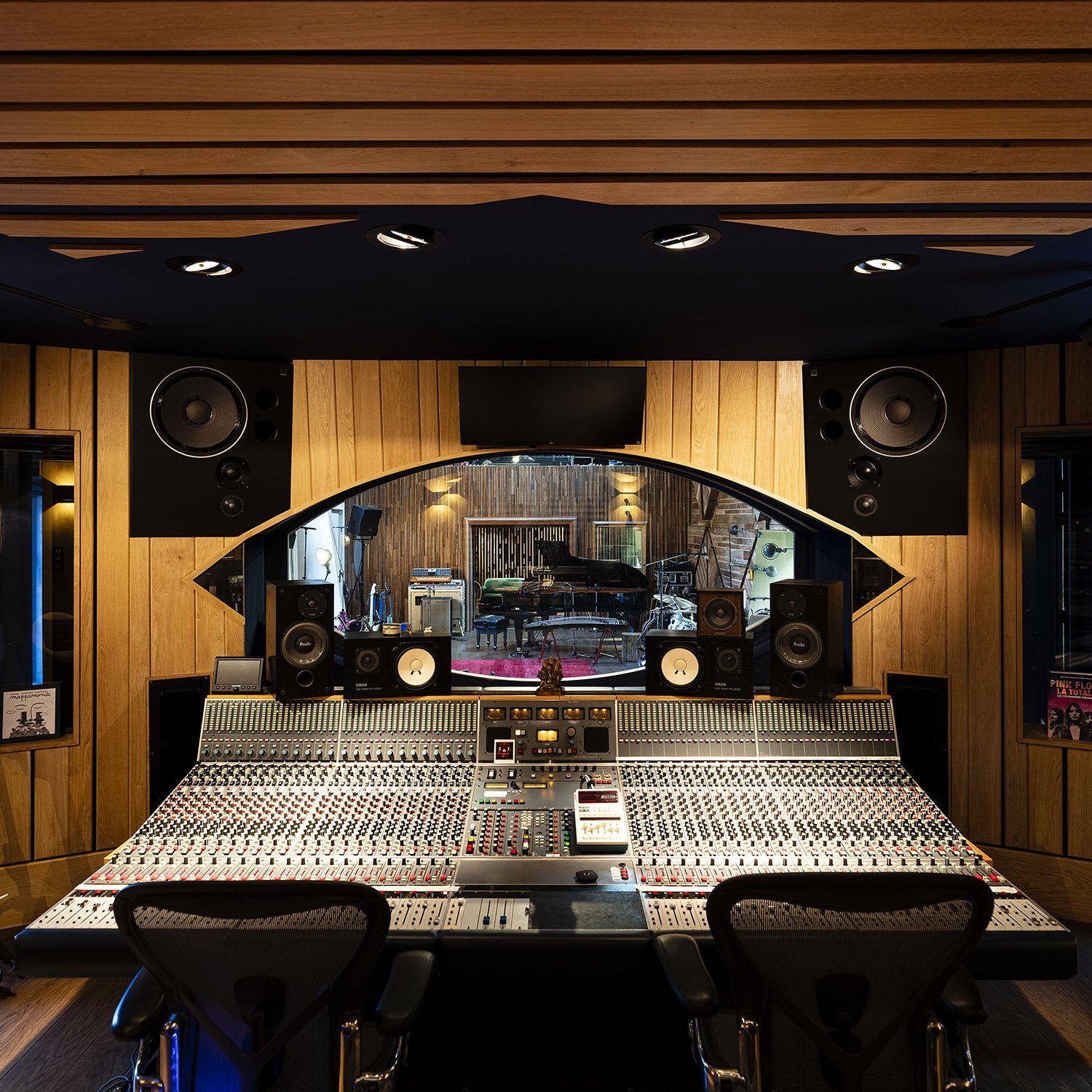 A professional recording studio with a large mixing console, studio monitors, and acoustic wooden wall panels, with a view into a room containing a grand piano and musical instruments.