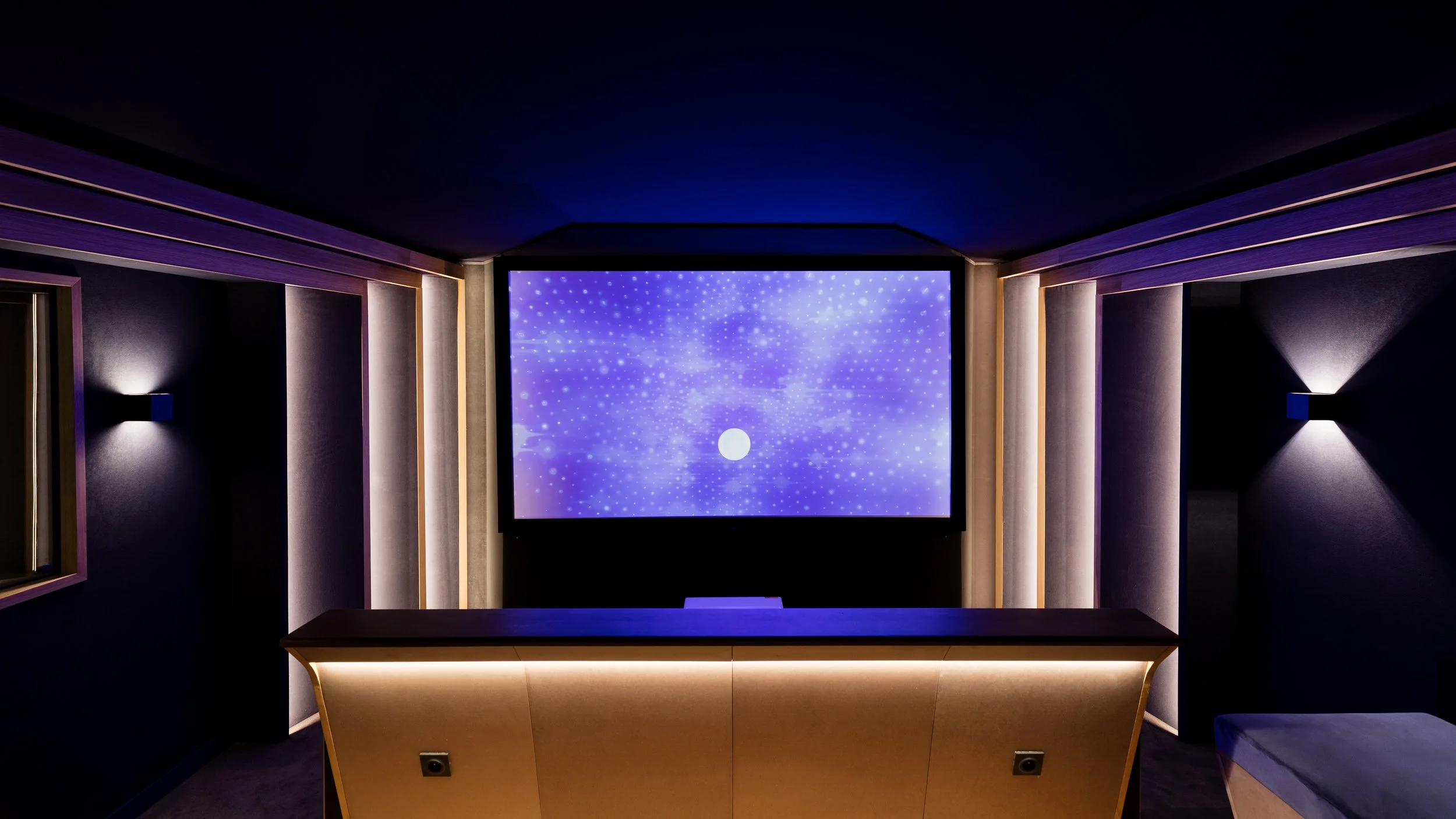 Salle de projection avec écran géant affichant des images de galaxies et d'étoiles, mobilier en bois clair, murs sombres avec éclairages subtils.