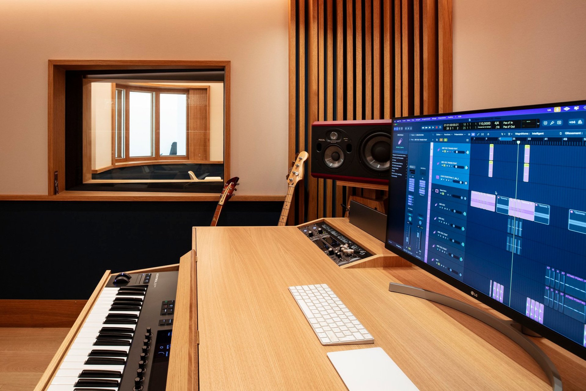 Studio d'enregistrement musical avec un clavier MIDI, un ordinateur avec logiciel de production musicale, des haut-parleurs et deux guitares sur des supports.