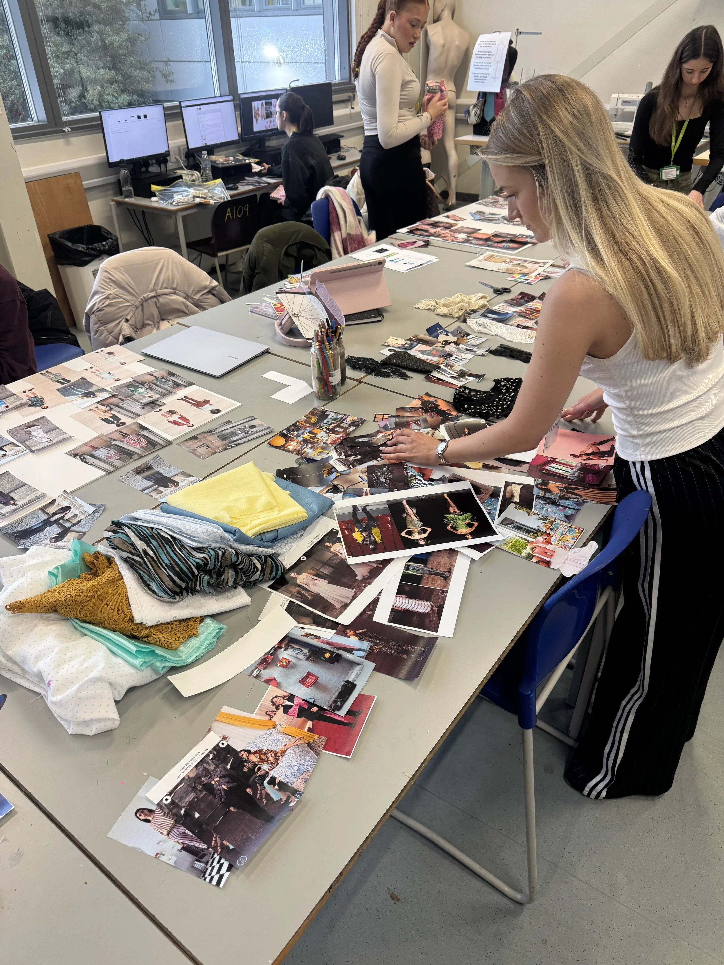 People working on a fashion photoshoot planning for a magazine, with magazines, clothing, and accessories on a table in a studio or office setting.