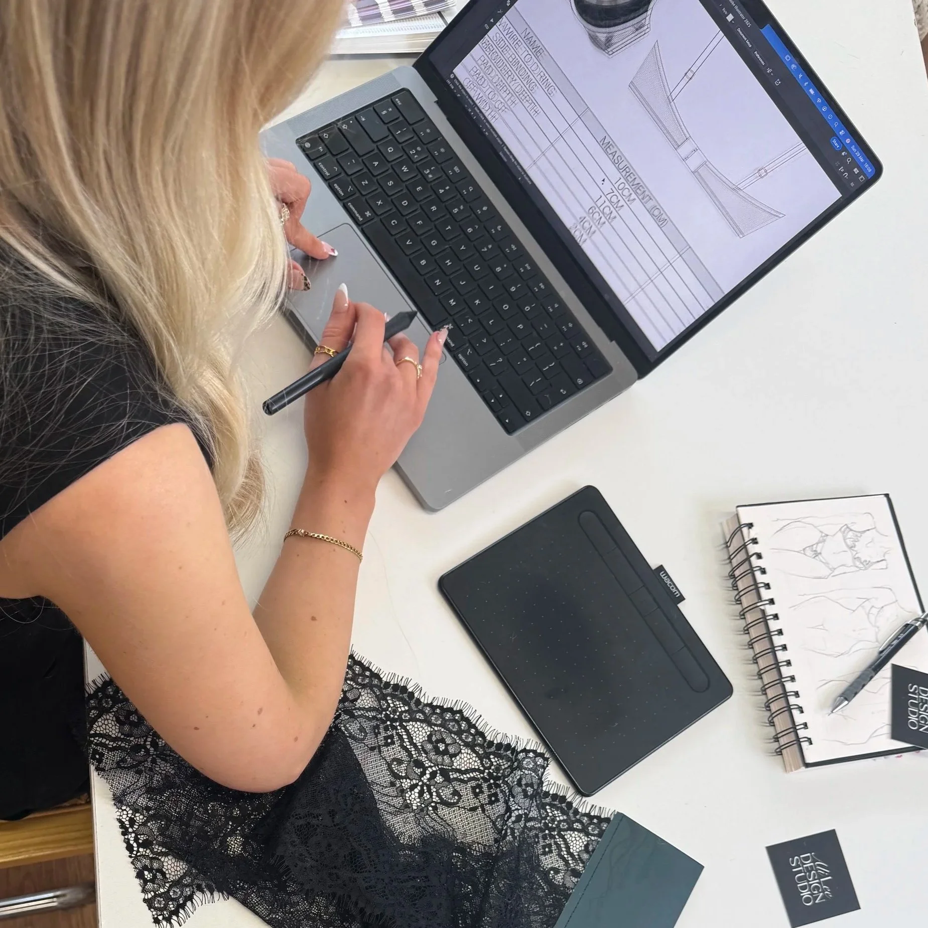 A woman with blonde hair and rings on her fingers is working on a laptop. The screen displays a technical drawing or blueprint with measurements and annotations. There are a black graphics tablet, a sketchbook with a fashion sketch, and business cards on the desk.