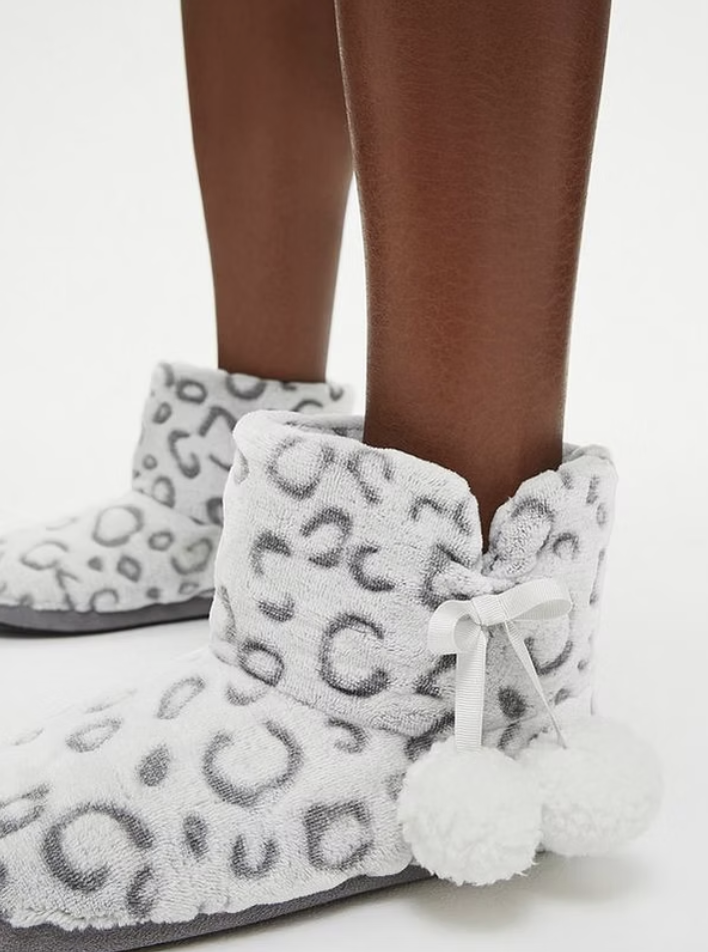 Close-up of legs wearing leopard print fluffy slippers with bows and pom-poms, against a plain white background.