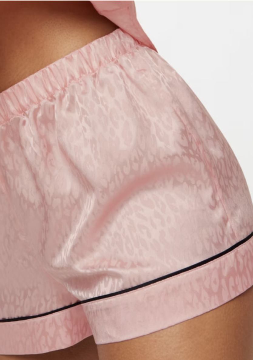Close-up of a person wearing pink satin pajama shorts with a leopard print pattern and a black piping detail.