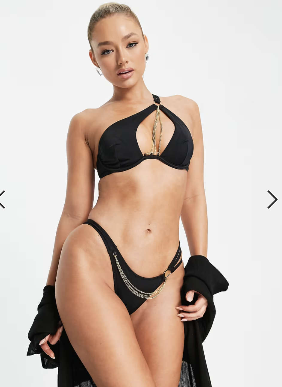 A woman wearing black lingerie with gold chains and a revealing top, holding a black garment, posing against a plain white background.