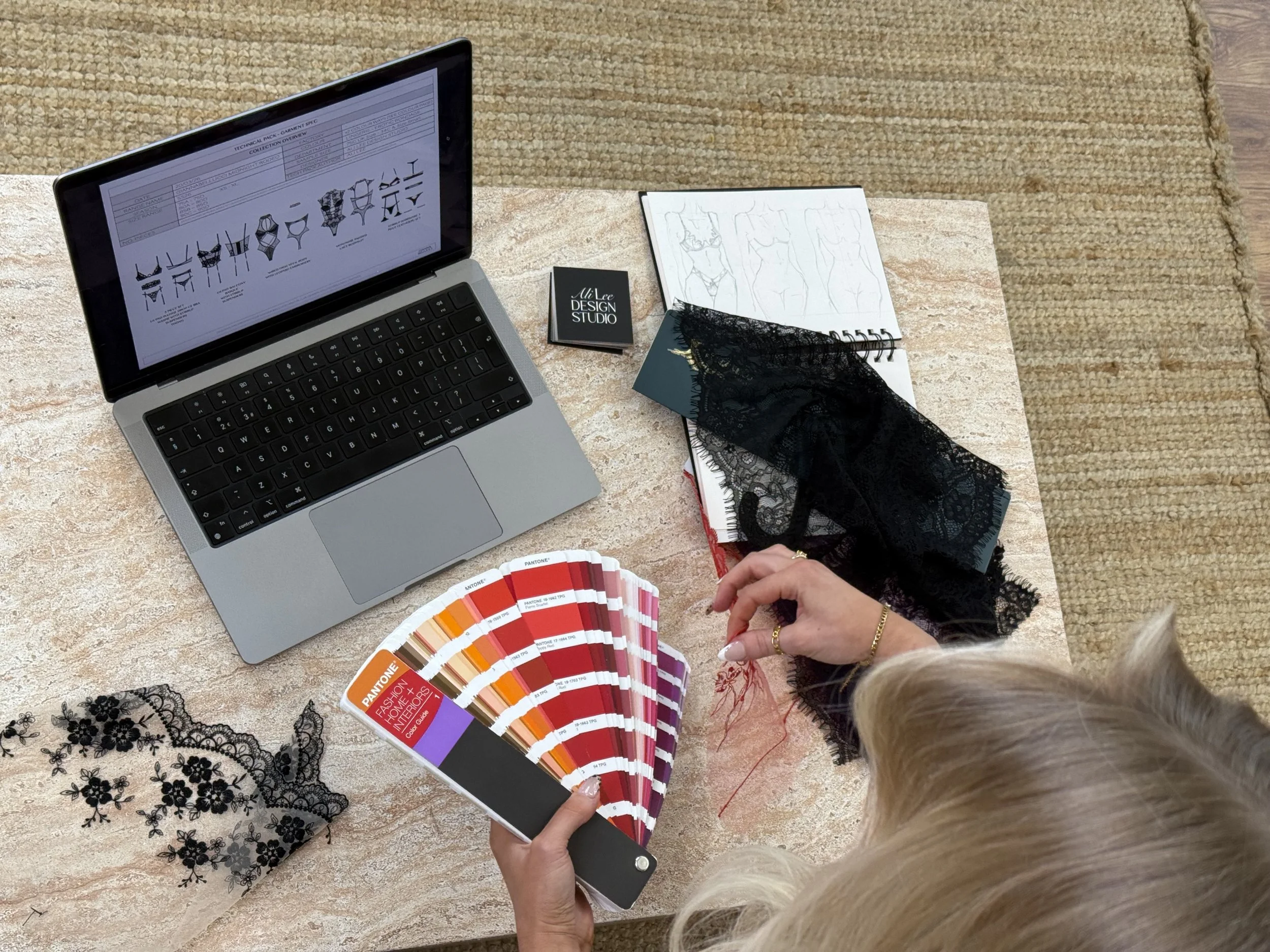 A person holding a Pantone color guide fan deck in front of a sewing pattern book, with a laptop and fabric swatches on a textured surface.