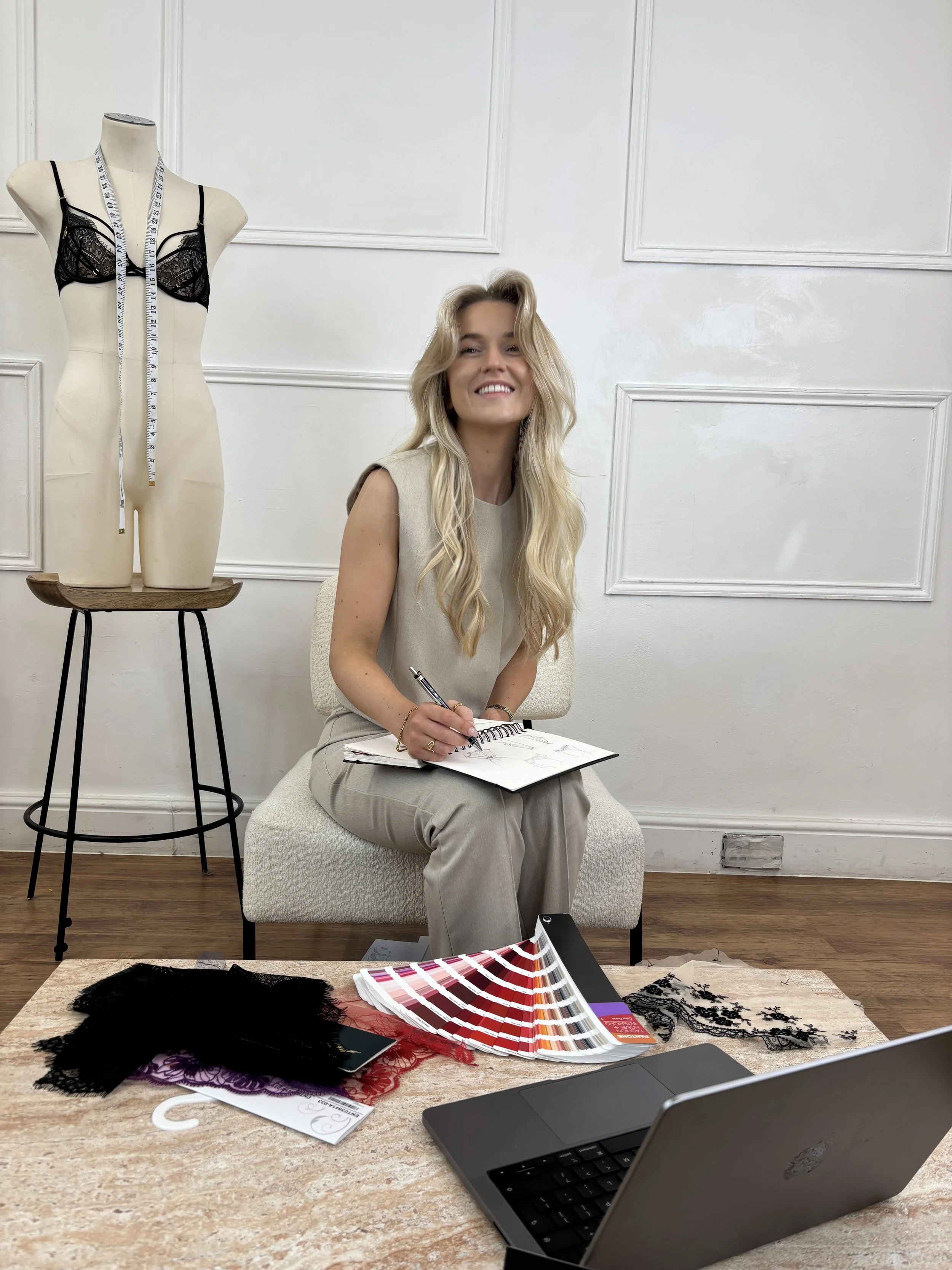 A woman sitting on a beige chair in a design studio, sketching on a notepad. A mannequin with a measuring tape is nearby. A table displays fabric swatches, lace trims, and color swatches. A laptop is open on the table.