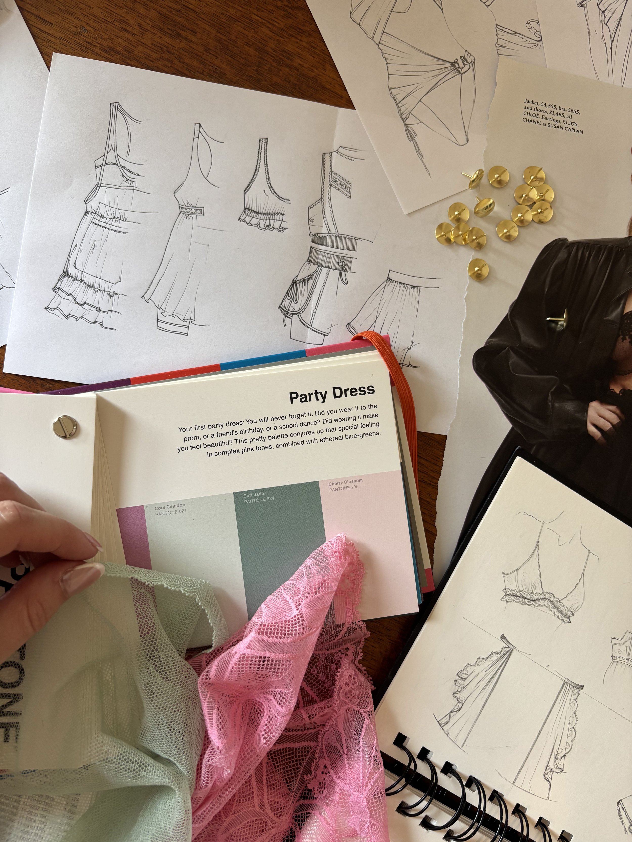 Sketches of party dresses on paper, fabric swatches in pink and beige, gold buttons, a photo of a woman in a black leather jacket, and a color palette sample.