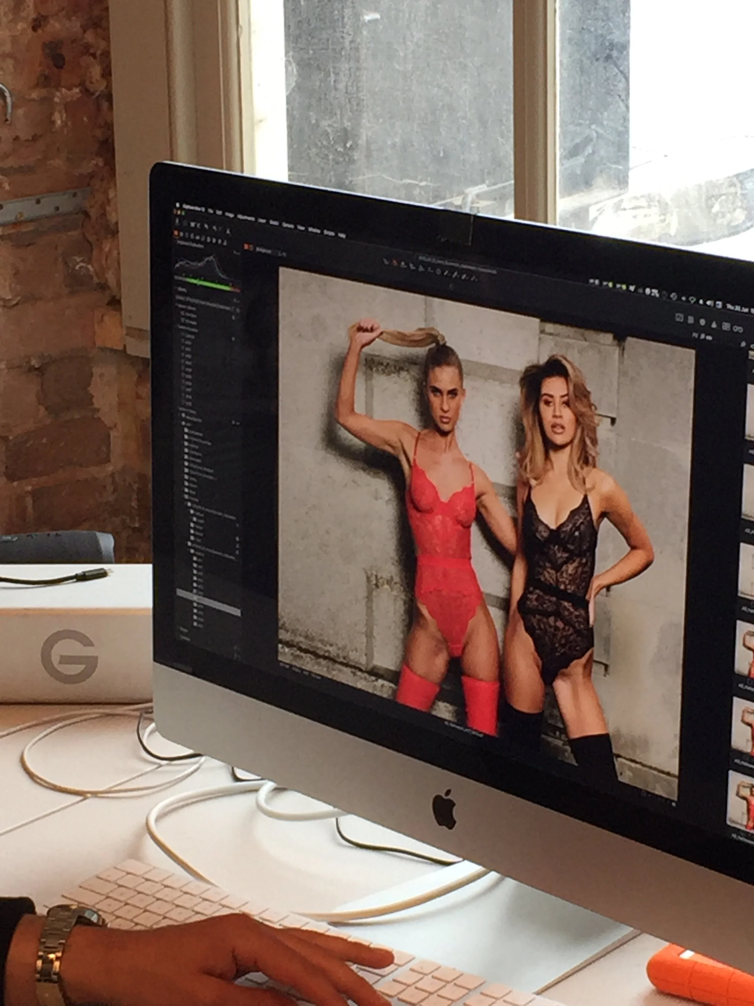 Computer monitor displaying a photoshoot of two women wearing lace lingerie and thigh-high stockings, with one woman holding her hair up.