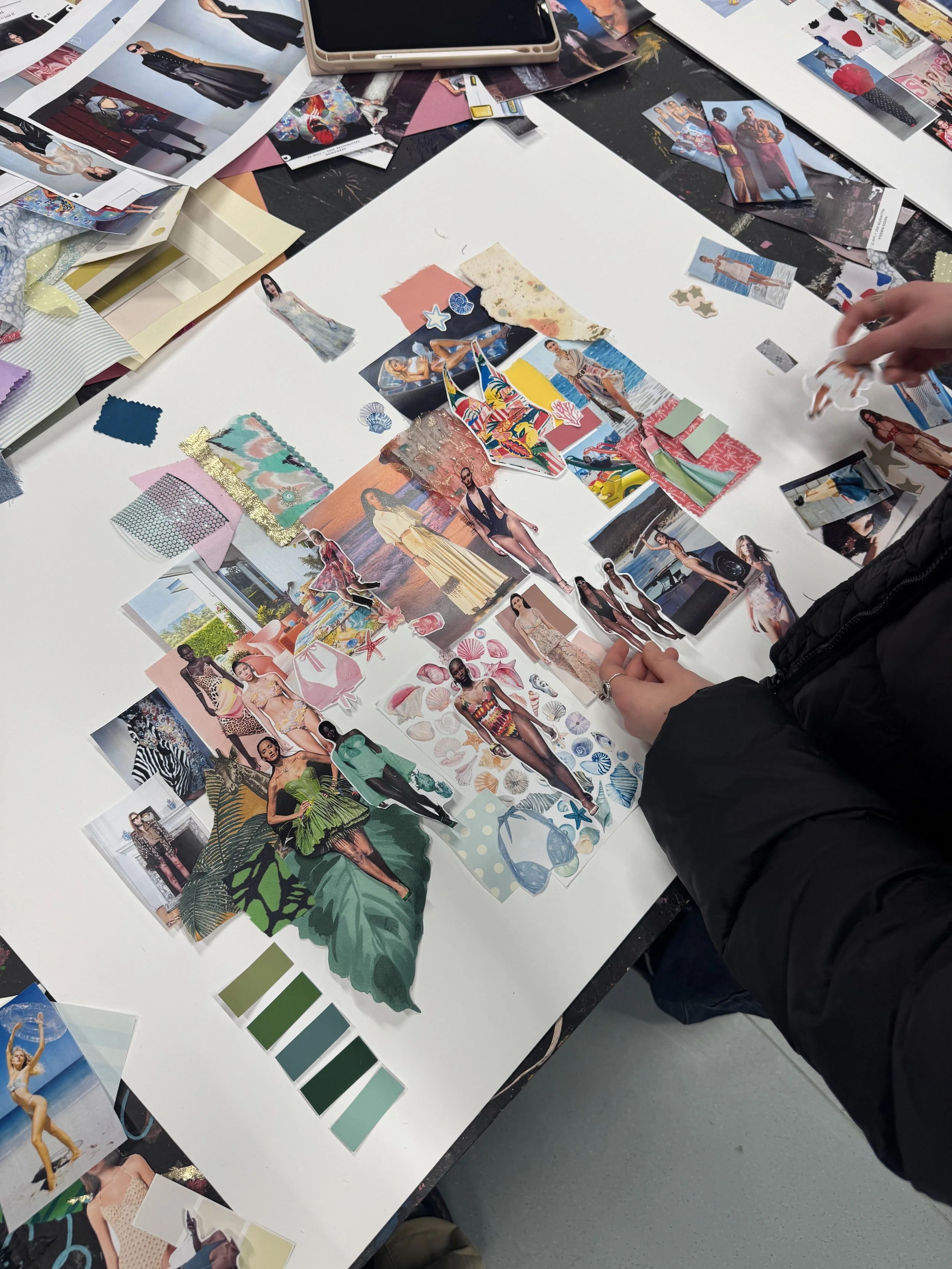 Table with fashion photos, fabric swatches, and design materials spread out. A person's hand is reaching across the table, working on a fashion storyboard or collage.