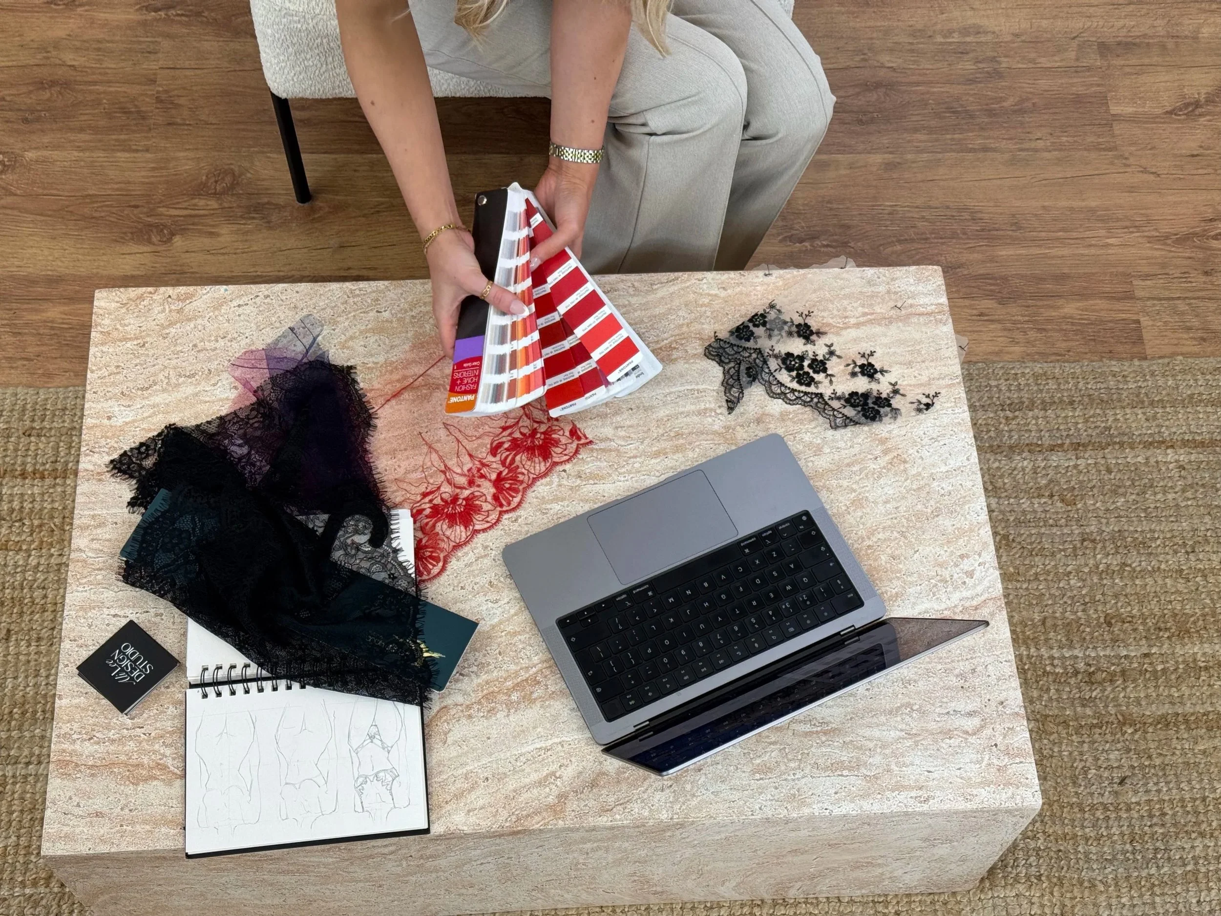 A person sitting at a wooden table displaying fabric swatches in red, black, and purple, alongside a sketchbook with fashion design sketches, a laptop, and other fashion design materials.