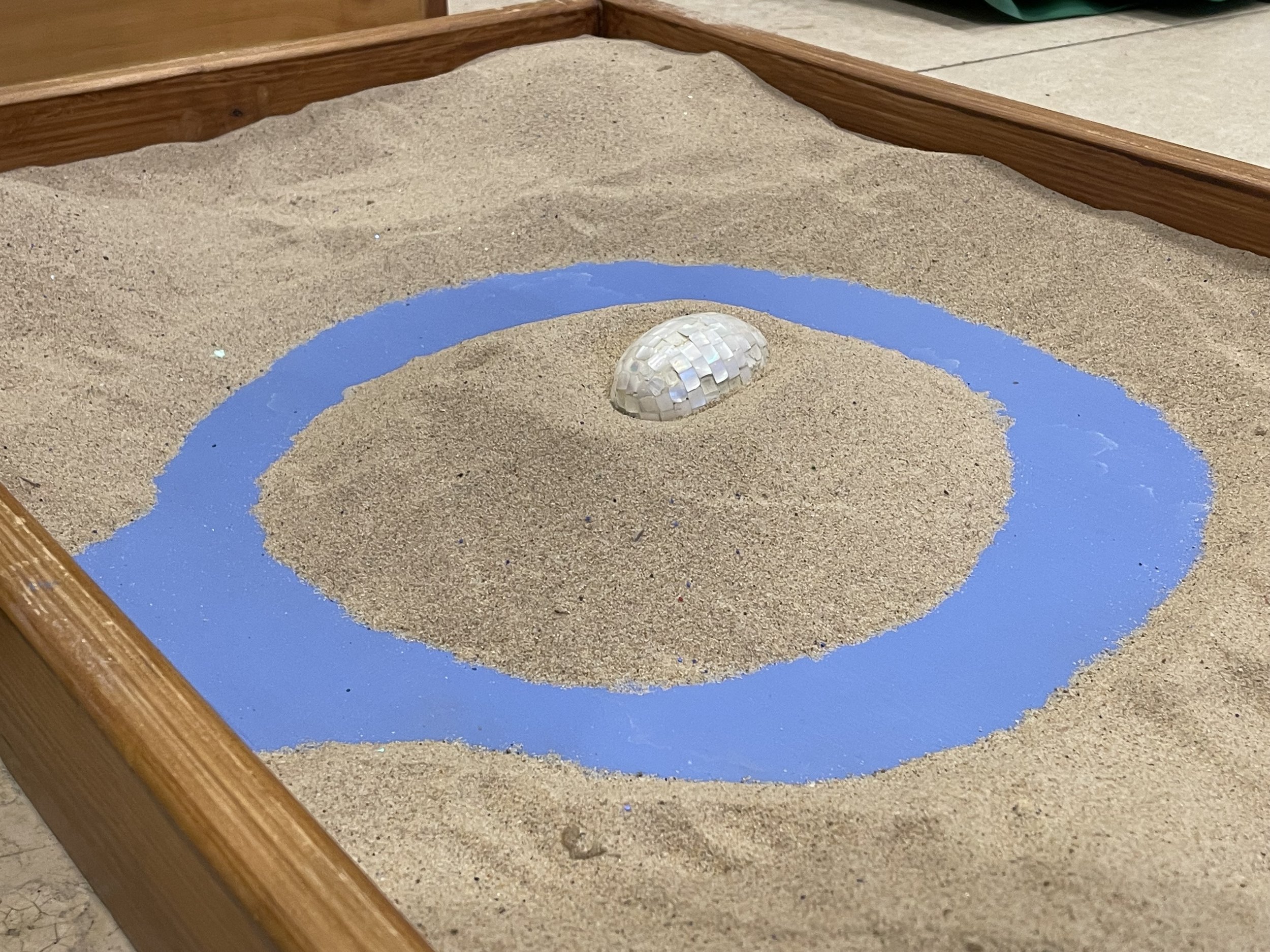 A sandbox with a blue moat dug in the sand and a mosaic egg on the island in the middle.
