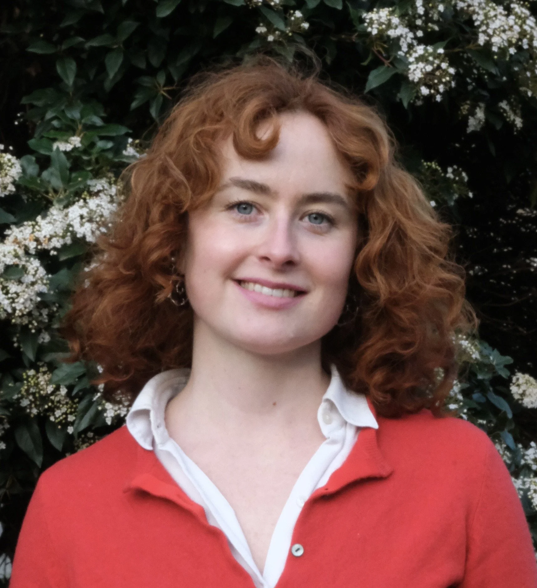A woman with curly red hair smiling outdoors, wearing a red sweater over a white collared shirt, with greenery and white flowers in the background.