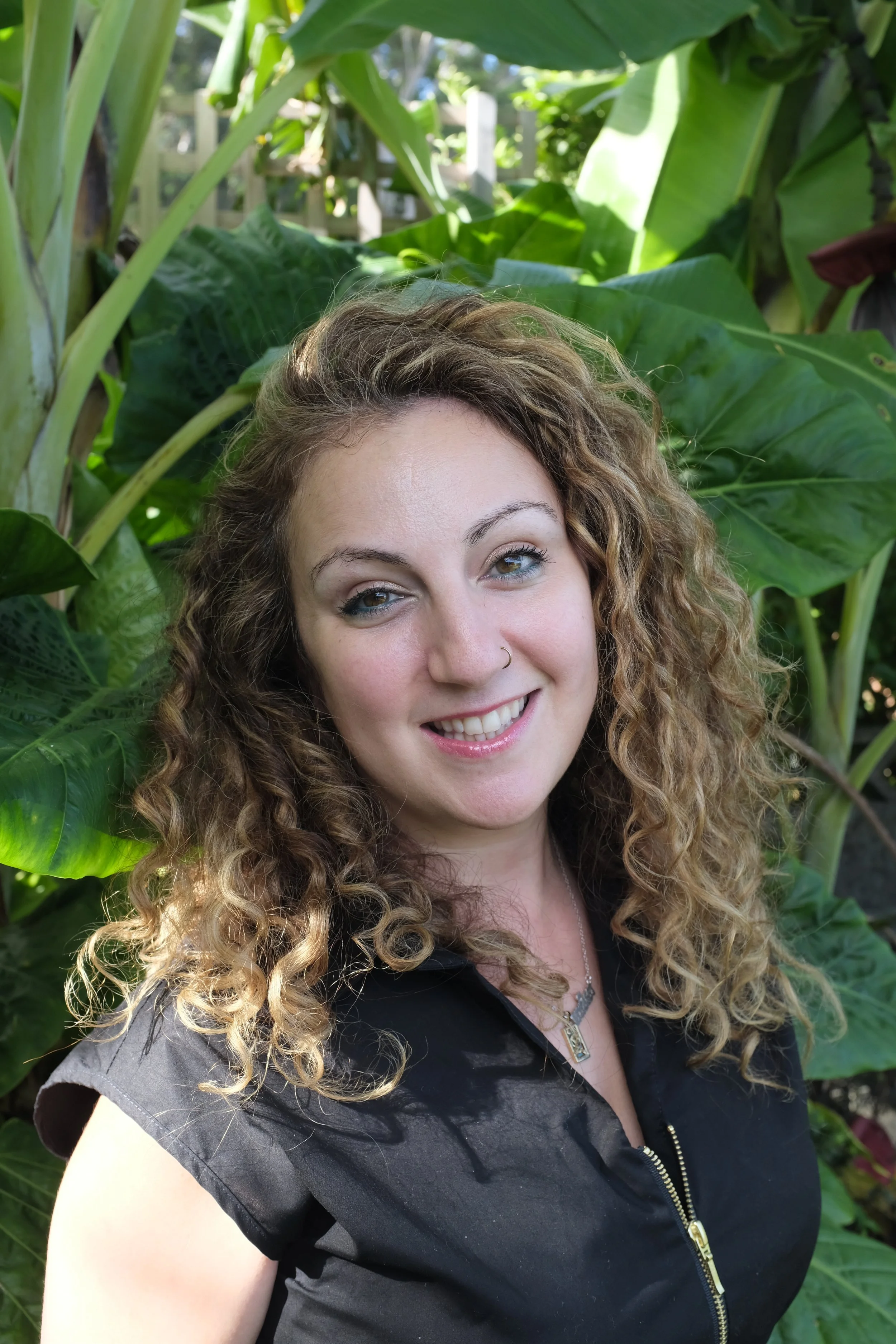 A  woman with curly hair smiling, wearing a black top with a zipper, standing among large green tropical leaves.