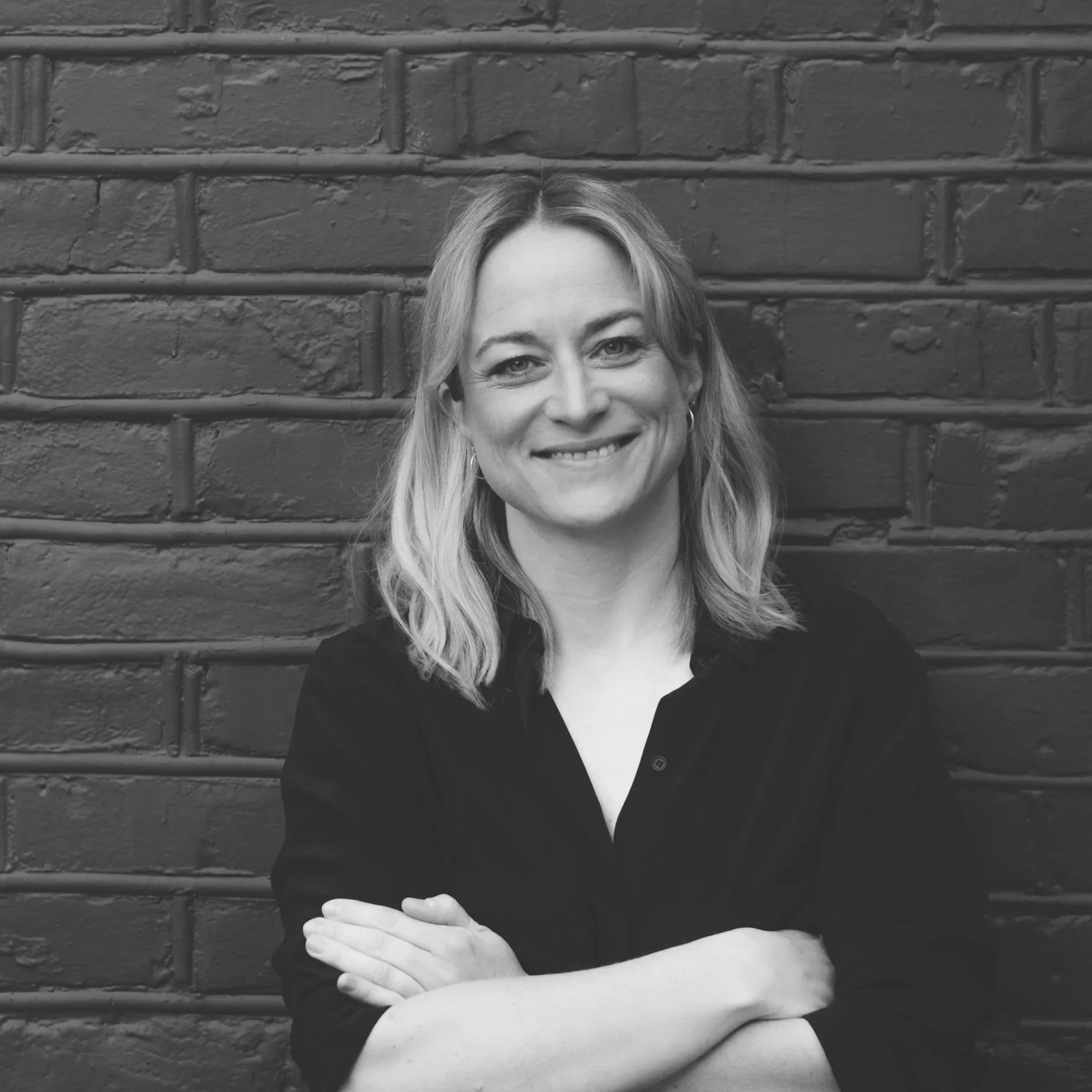 Portrett av en smilende kvinne med blondt hår, kledd i svart, foran en murvegg.