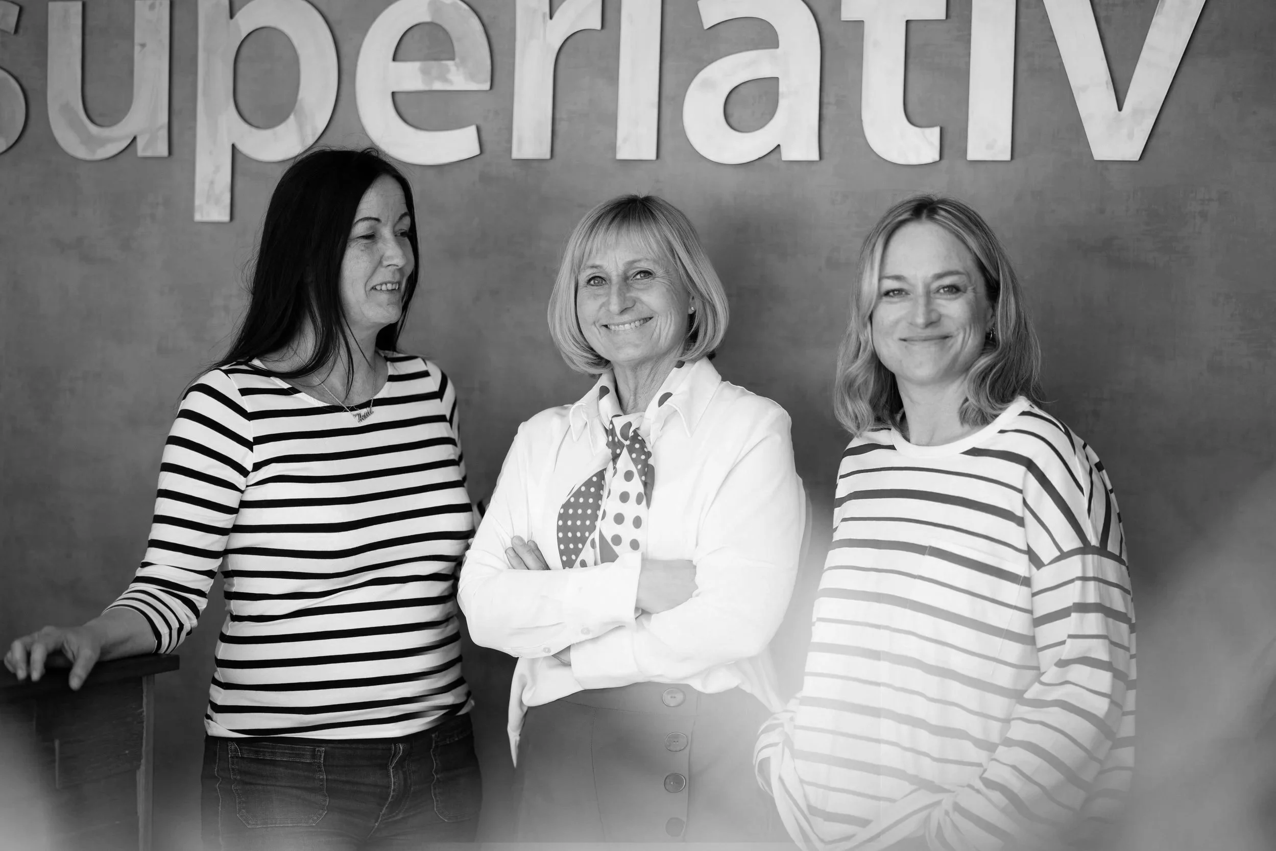 Tre kvinner som står foran en vegg med store bokstaver. To av dem smiler og en ser ut til å snakke. Alt er i svart-hvitt.