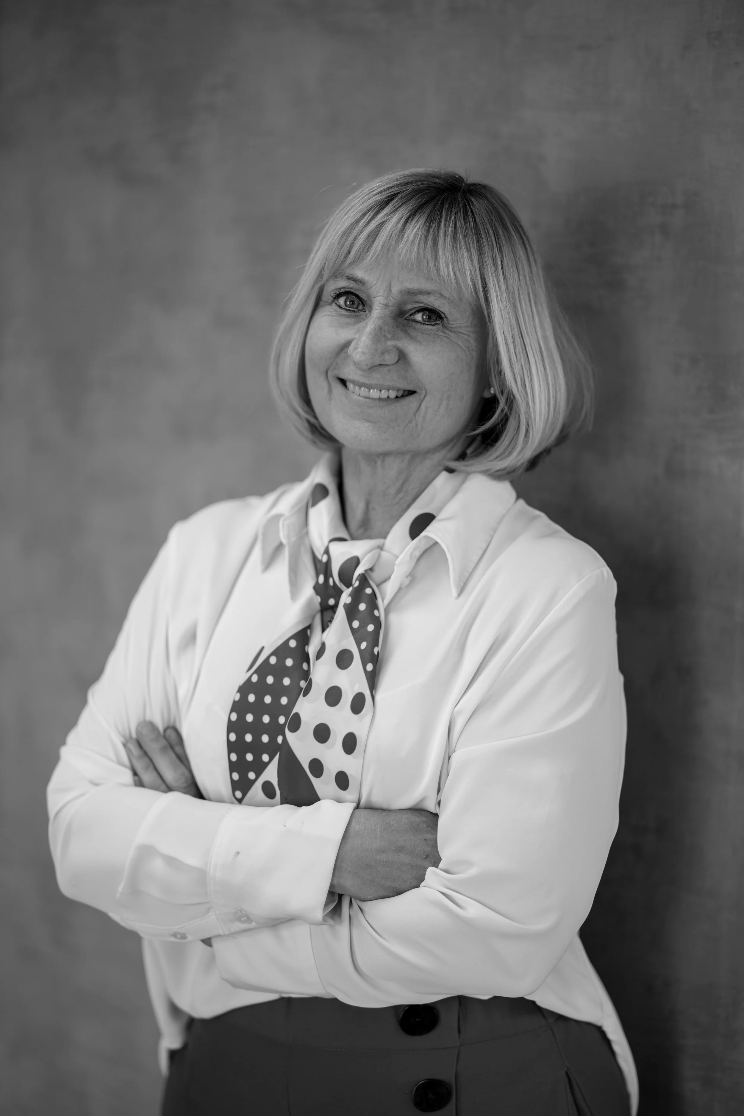 En eldre kvinne med kort blond hår smiler mot kamera, kledd i hvit skjorte og en stripete skjerf, står foran en enkel vegg.