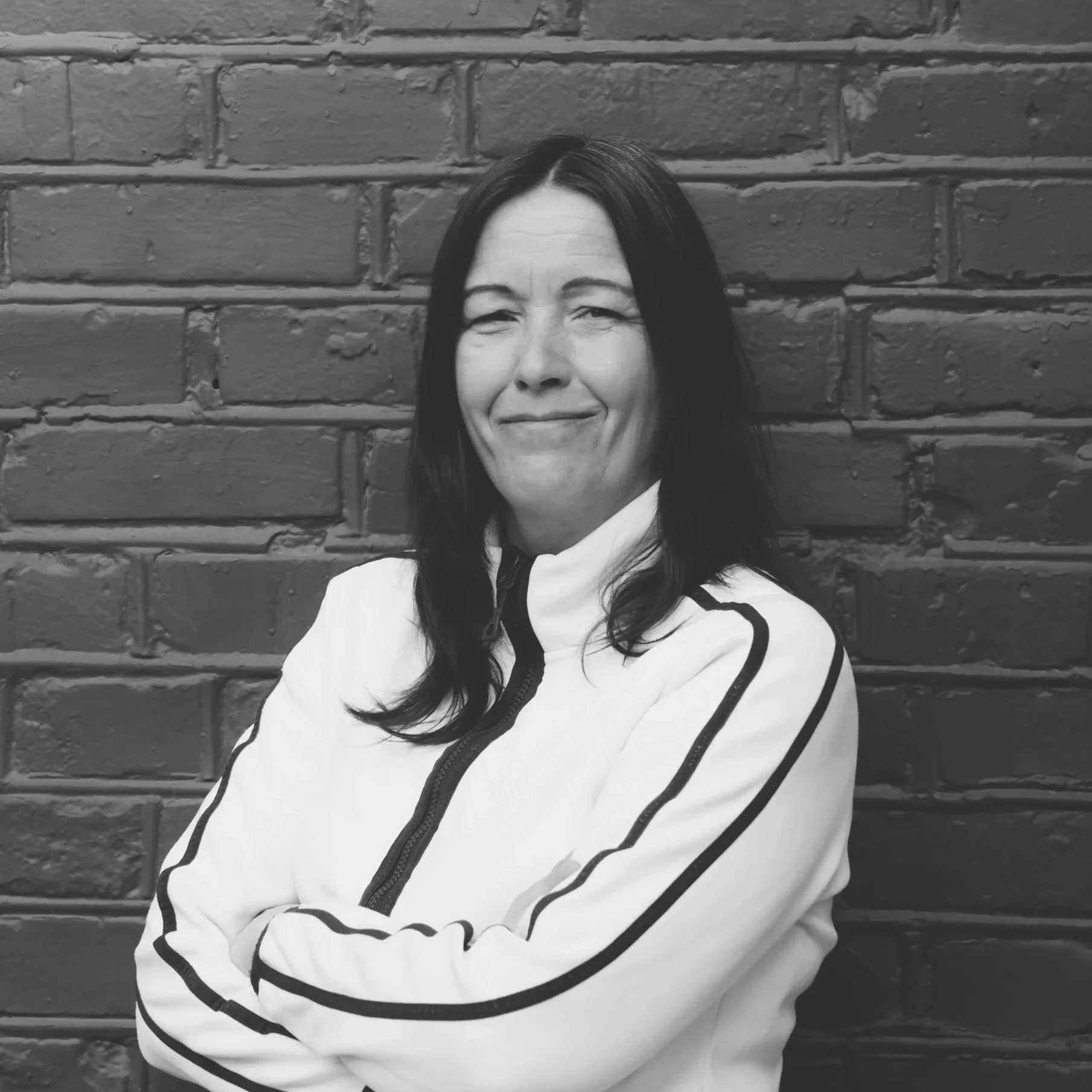 Sort-hvitt portrett av en kvinne med mørkt hår som smiler, står foran en mur av murstein.